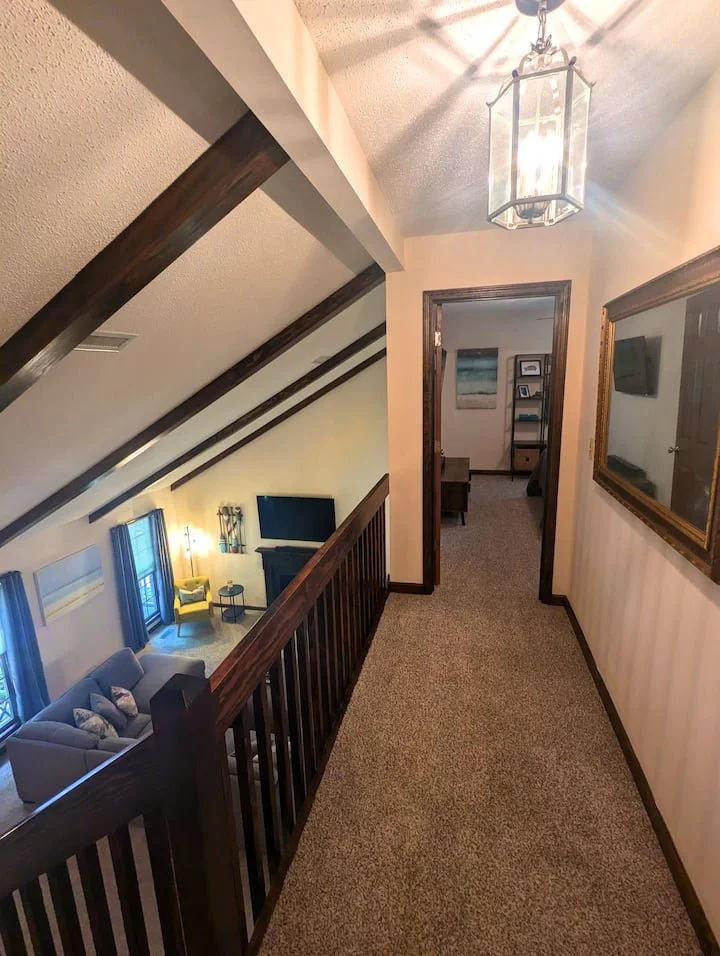 Hallway with carpeted floor, with a view into a living room below. The hallway has wood trim and a mirror on the right wall. There is a ceiling light fixture hanging from the ceiling.