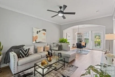 Bright living room with gray walls, white trim, large sectional sofa with decorative pillows, glass coffee table, indoor plants, and a ceiling fan. French doors lead to a balcony with outdoor seating.
