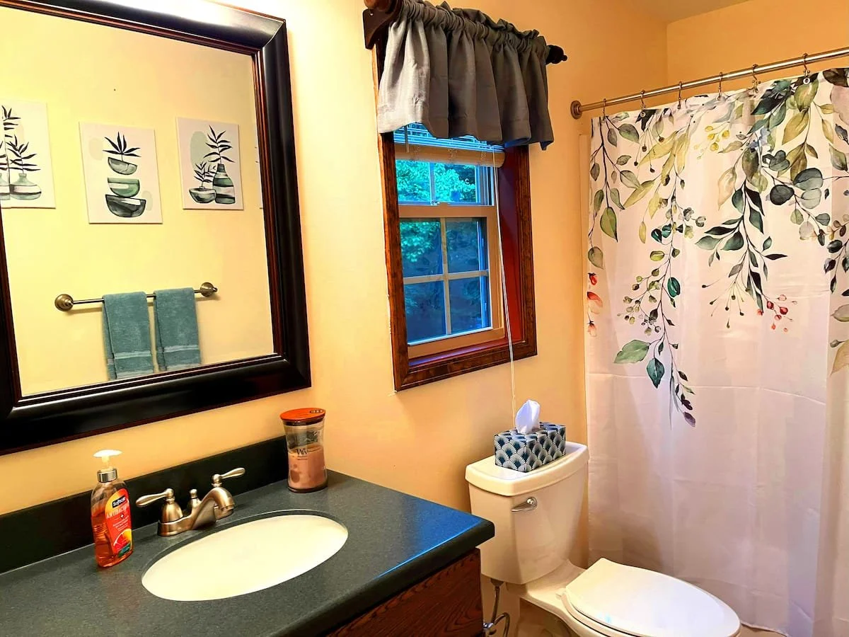 A small bathroom with a mirror, a sink with soap, a window with a curtain, a toilet with a tissue box on top, and a bathtub with a floral shower curtain.