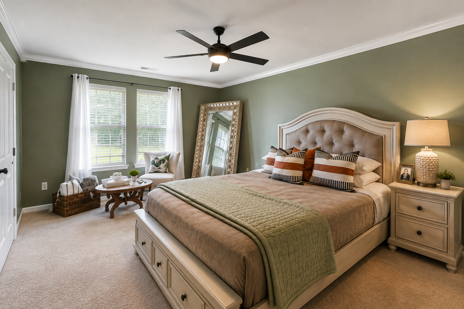 A bedroom with green walls, a large beige bed with pillows, and a white nightstand with a lamp and framed photo. There are two windows with white curtains, a full-length mirror leaning against the wall, and a sitting area with a small round table, an armchair, and a basket with blankets.