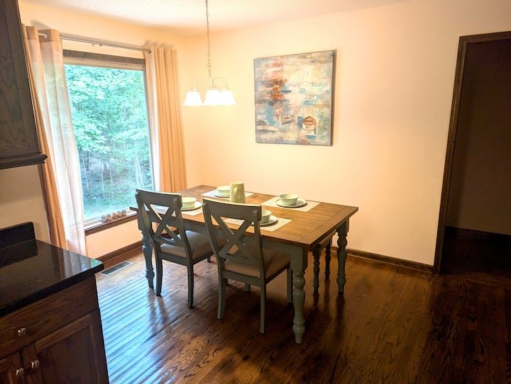 Dining room with a wooden table set for four, featuring plates, bowls, and cups. Four chairs are arranged around the table. A large window with curtains is on the left, letting in natural light and showing greenery outside. A painting is hung on the wall above the table, and a chandelier with two lights hangs overhead. The room has wood flooring and an open doorway to the right.