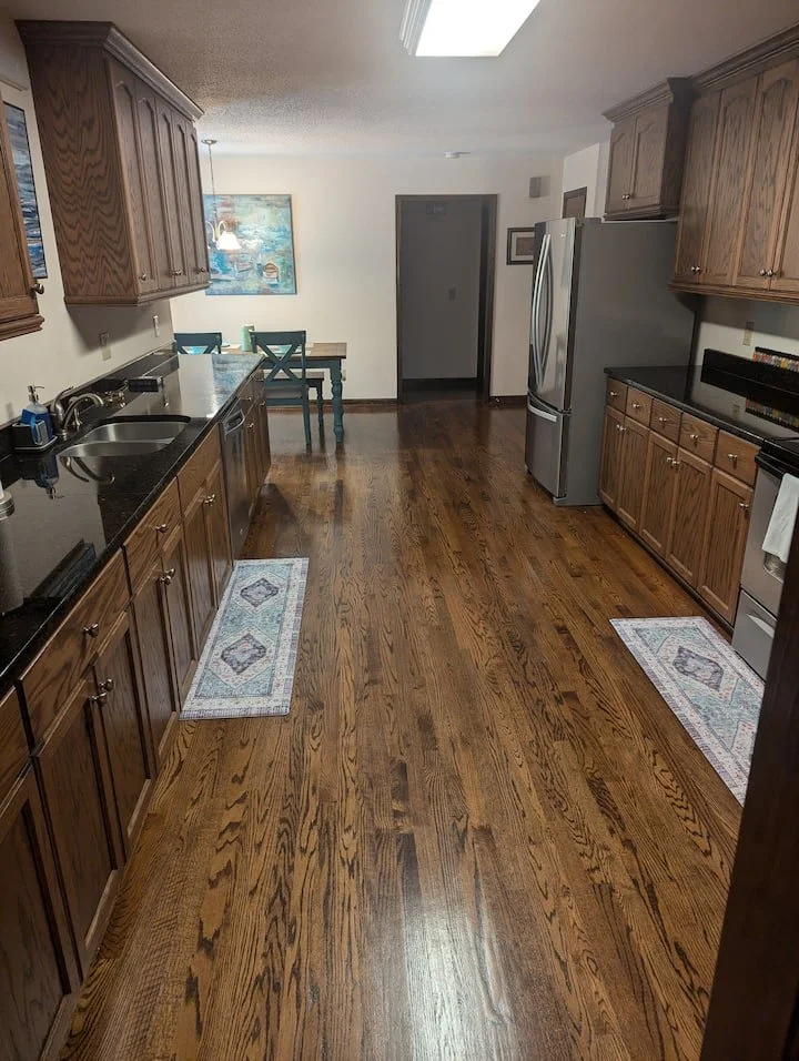 Empty kitchen with wooden cabinets, black countertops, stainless steel refrigerator, and a small dining table with chairs in the background.