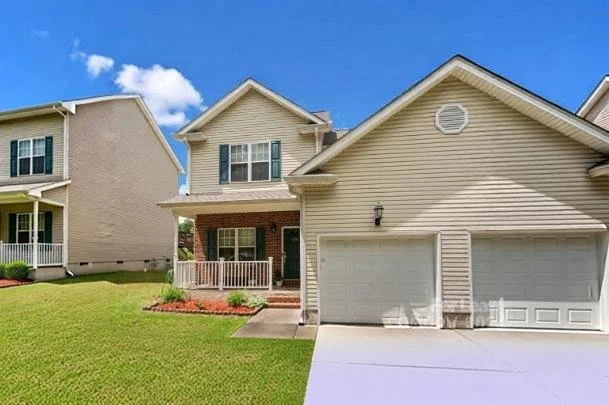 Front view of a suburban house with beige siding, attached garage, and well-maintained lawn under a partly cloudy sky.