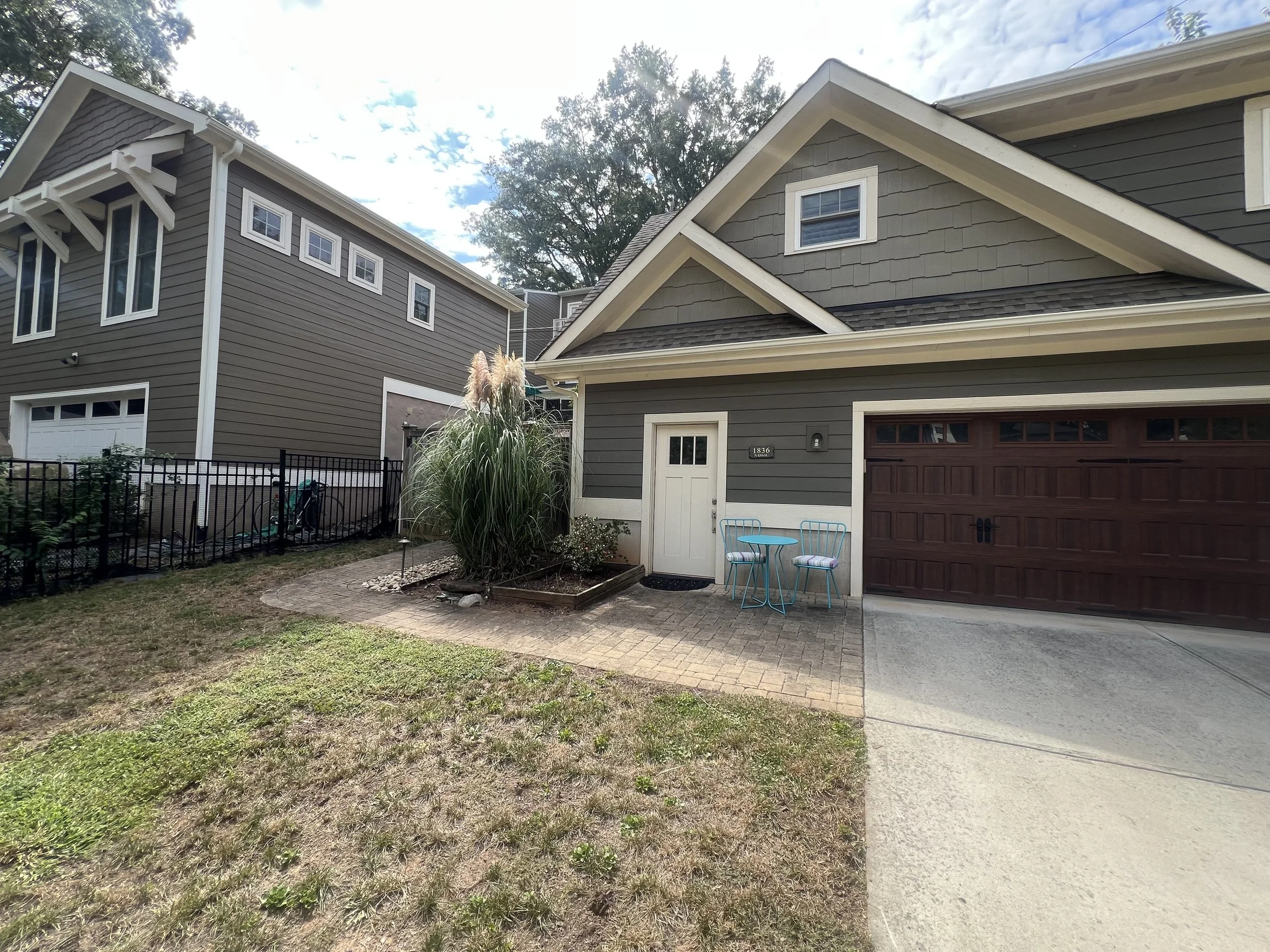 View of a residential house with garage, small patio area with two chairs and a table, tall ornamental grass in the front yard, lattice fencing, and a paved driveway under partly cloudy sky.