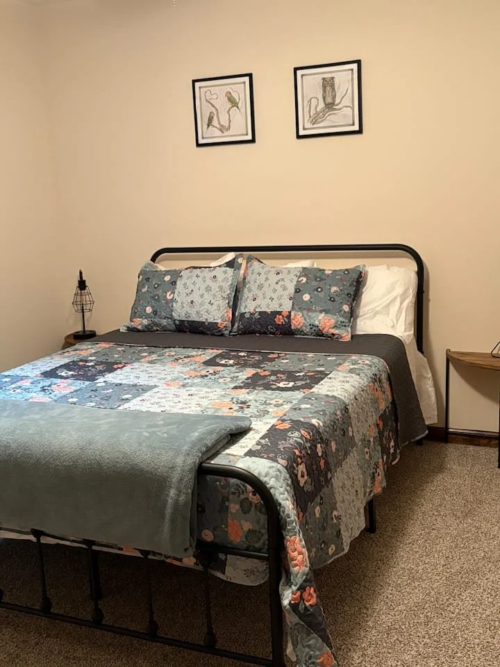 A neatly made bed with a floral quilt and matching pillows in a simple bedroom. There are framed line art pictures hanging on the cream-colored wall above the bed, a small black bedside lamp on the left, and a small wooden side table on the right. The room has a beige carpet.