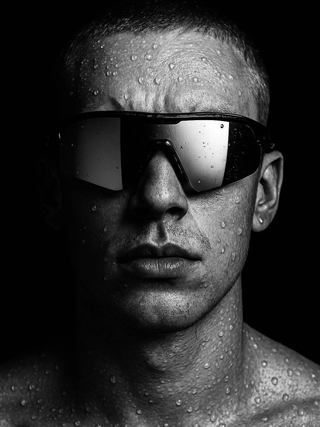 Black-and-white close-up photo of a man with water droplets on his face, wearing reflective sunglasses, against a dark background.