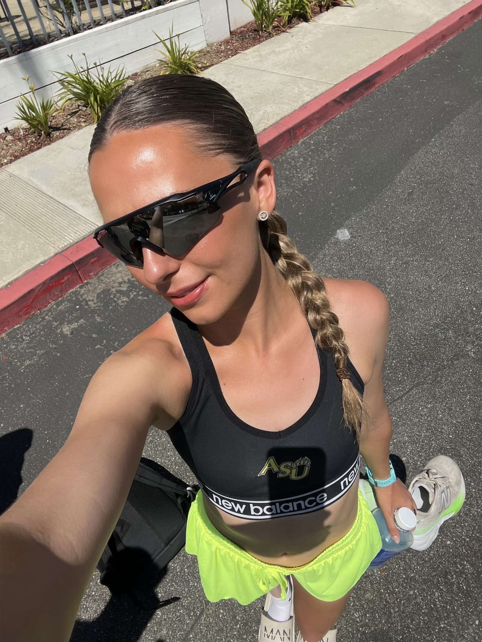 A woman taking a selfie outdoors, wearing large black sports sunglasses, a black sports bra with