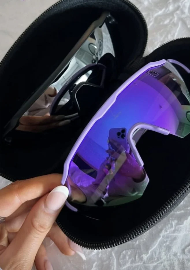 A person holding a pair of colorful, reflective sunglasses with purple, green, and blue iridescent lenses, in front of a black glasses case, with sunglasses inside the case and a smartphone reflected in the lenses.