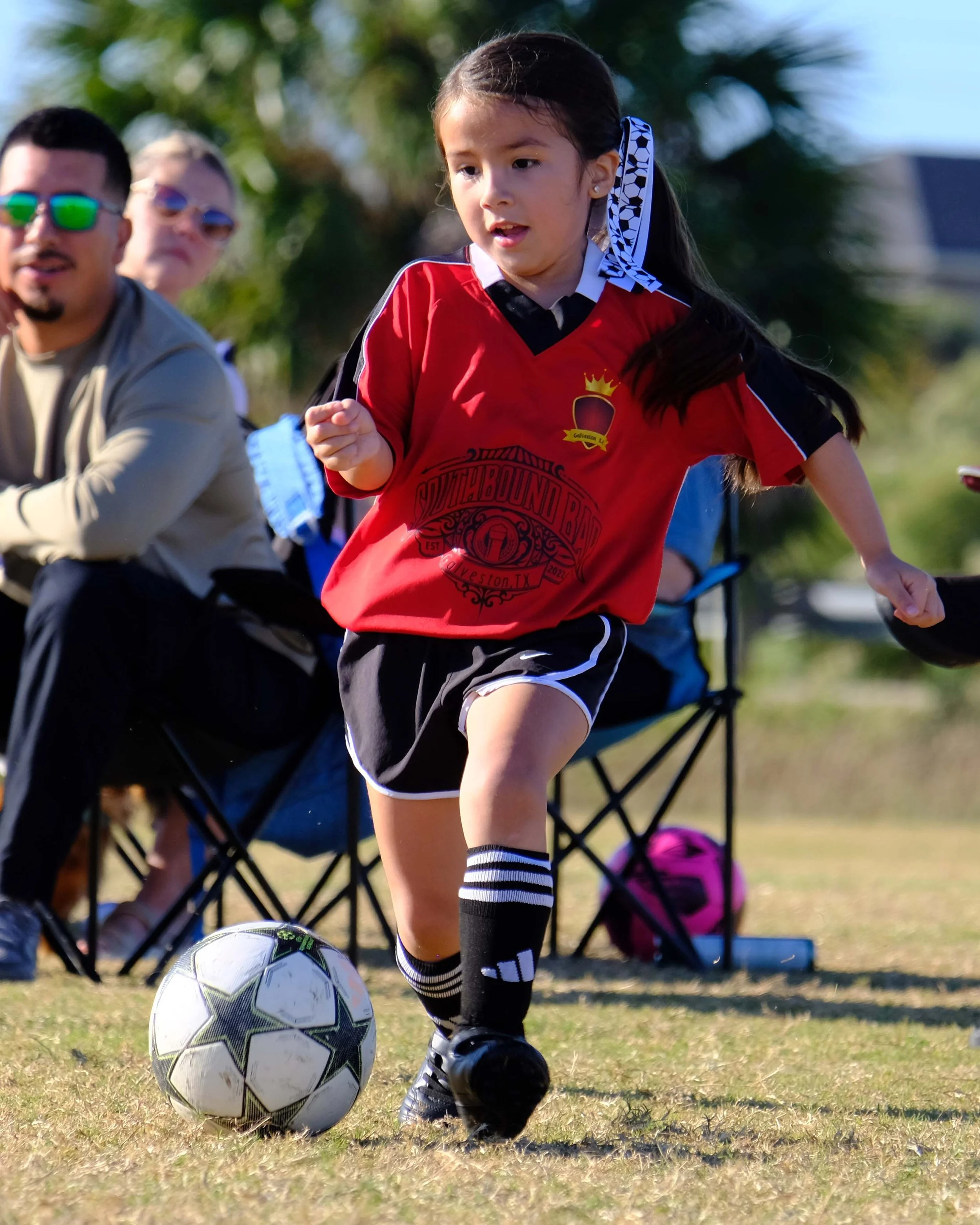 A young girl in a red soccer jersey and black shorts playing soccer on a grassy field, with a soccer ball in front of her, and a few people sitting on chairs in the background.