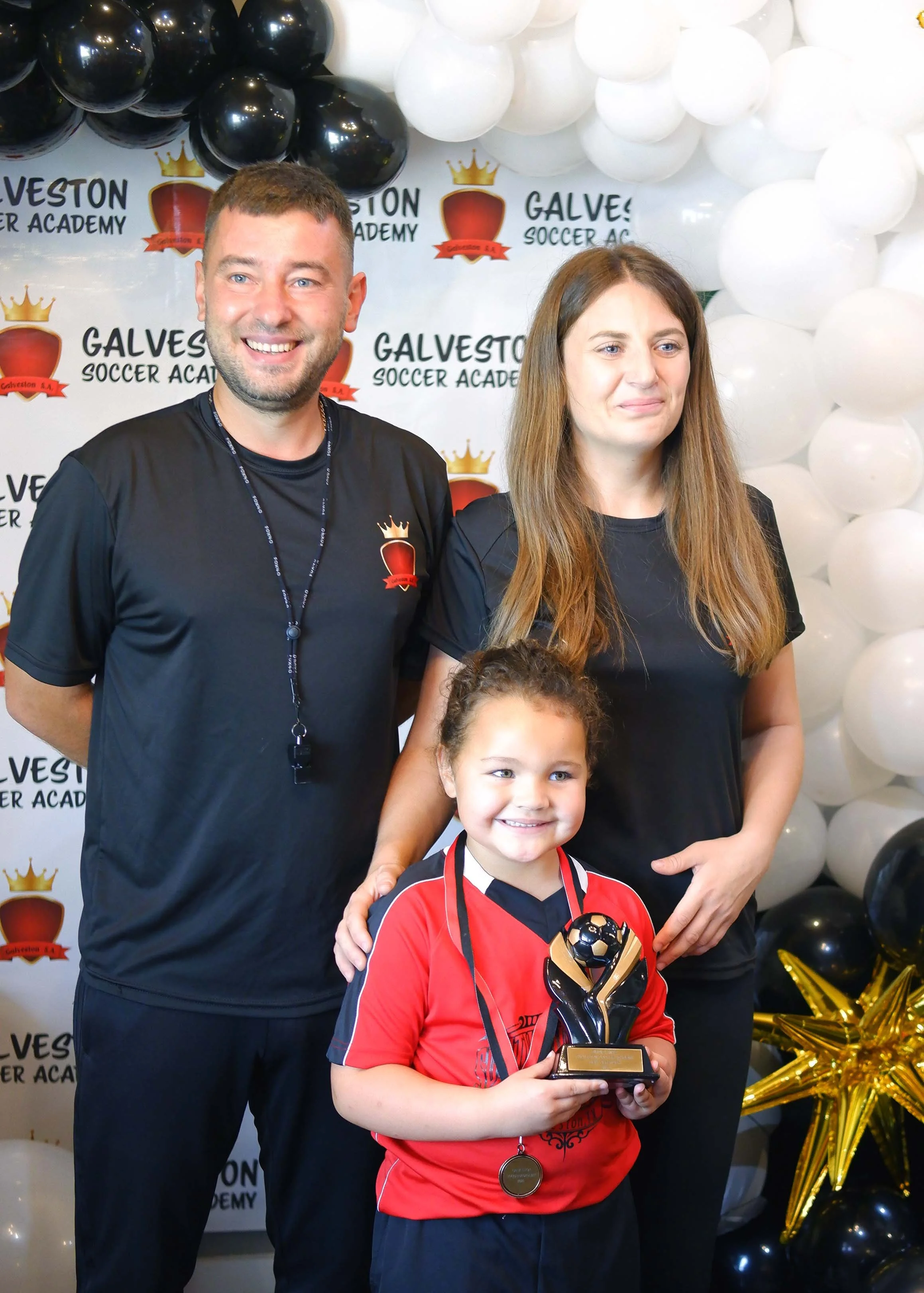 A young girl in a red sports jersey holding a trophy and medals, smiling at a celebration event, with a man and a woman standing behind her. The background features a wall with the logo of Galveston Soccer Academy and black, white, and gold balloons.