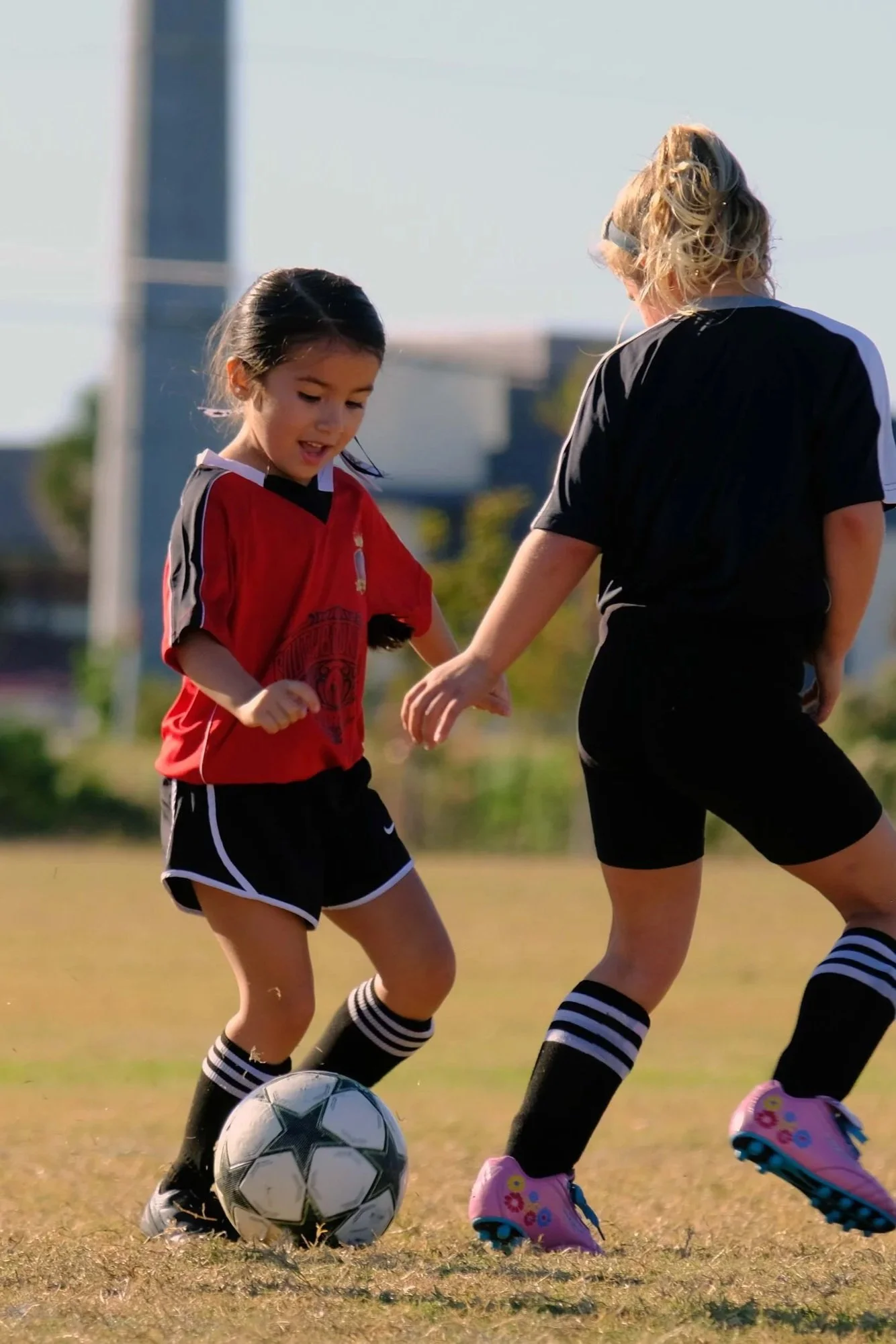A girl in a red soccer jersey and black shorts playing soccer with two women, one of whom has blonde hair and is wearing a black shirt with striped socks, on a grassy field on a sunny day.