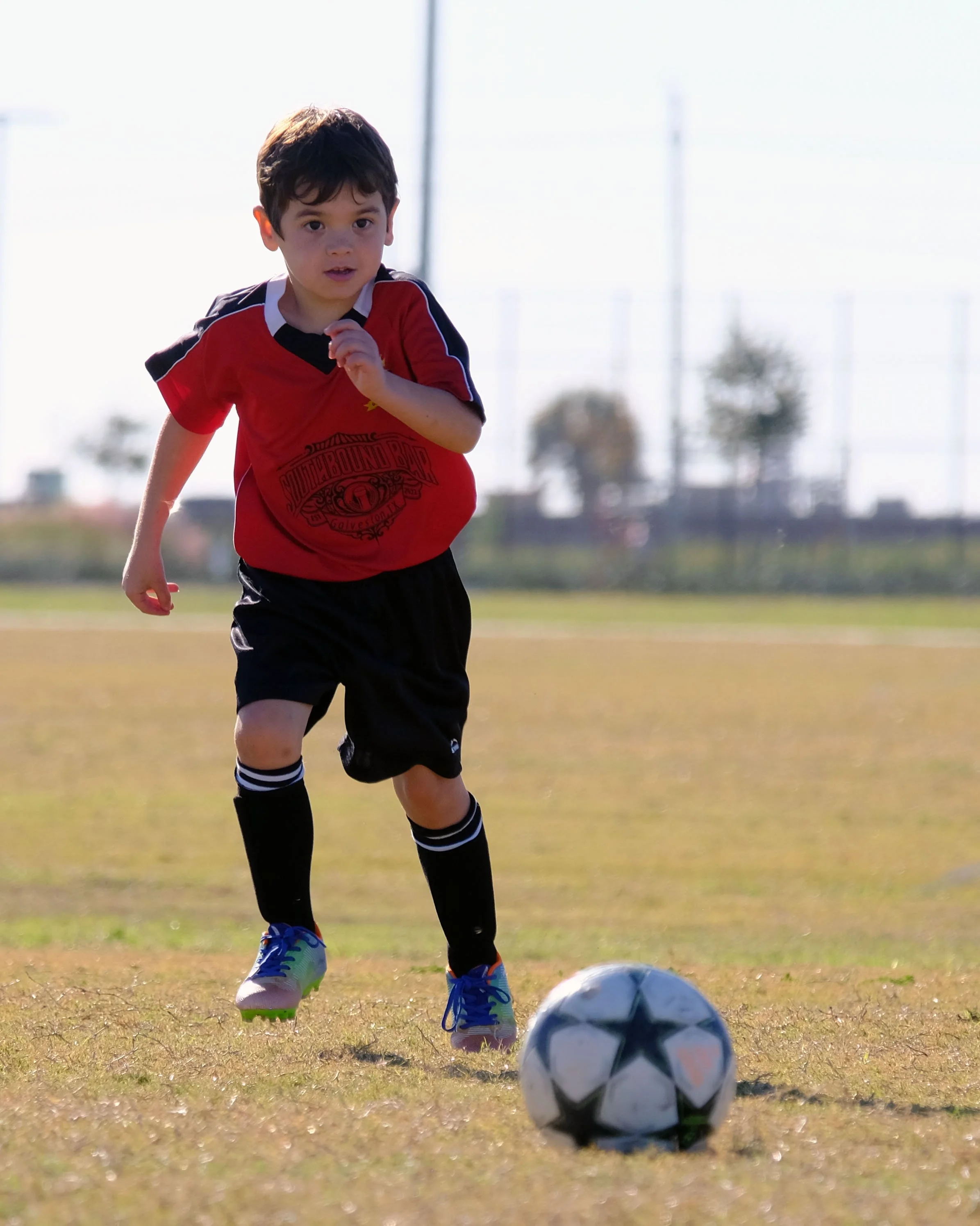 A young boy with brown hair wearing a red and black soccer uniform, black shorts, and blue and green cleats, is playing soccer on a field with a soccer ball nearby. The background is blurred with trees and power lines.