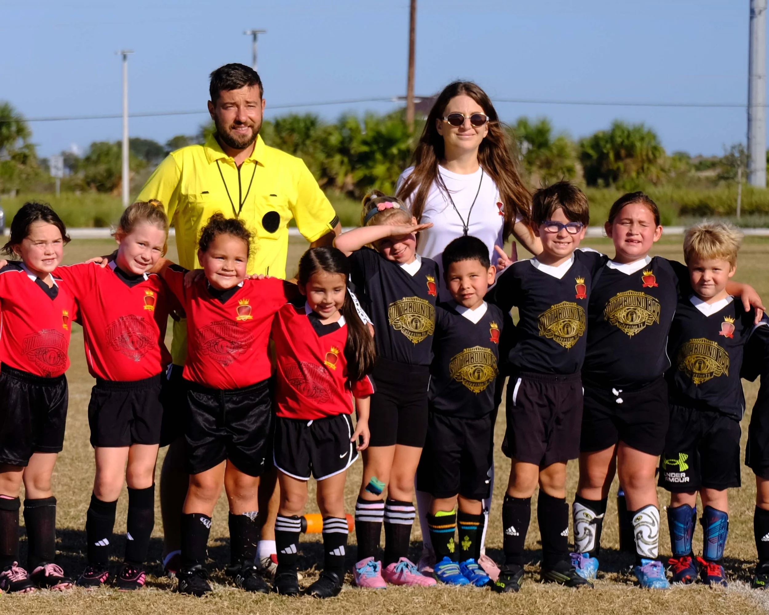 A group of young children and two adults posing together on a sports field, with clear blue sky, trees, and utility poles in the background. The kids are wearing athletic jerseys and shorts, some with logo graphics, and sneakers, with one child saluting and others smiling. The two adults, a man and a woman, stand behind the children.