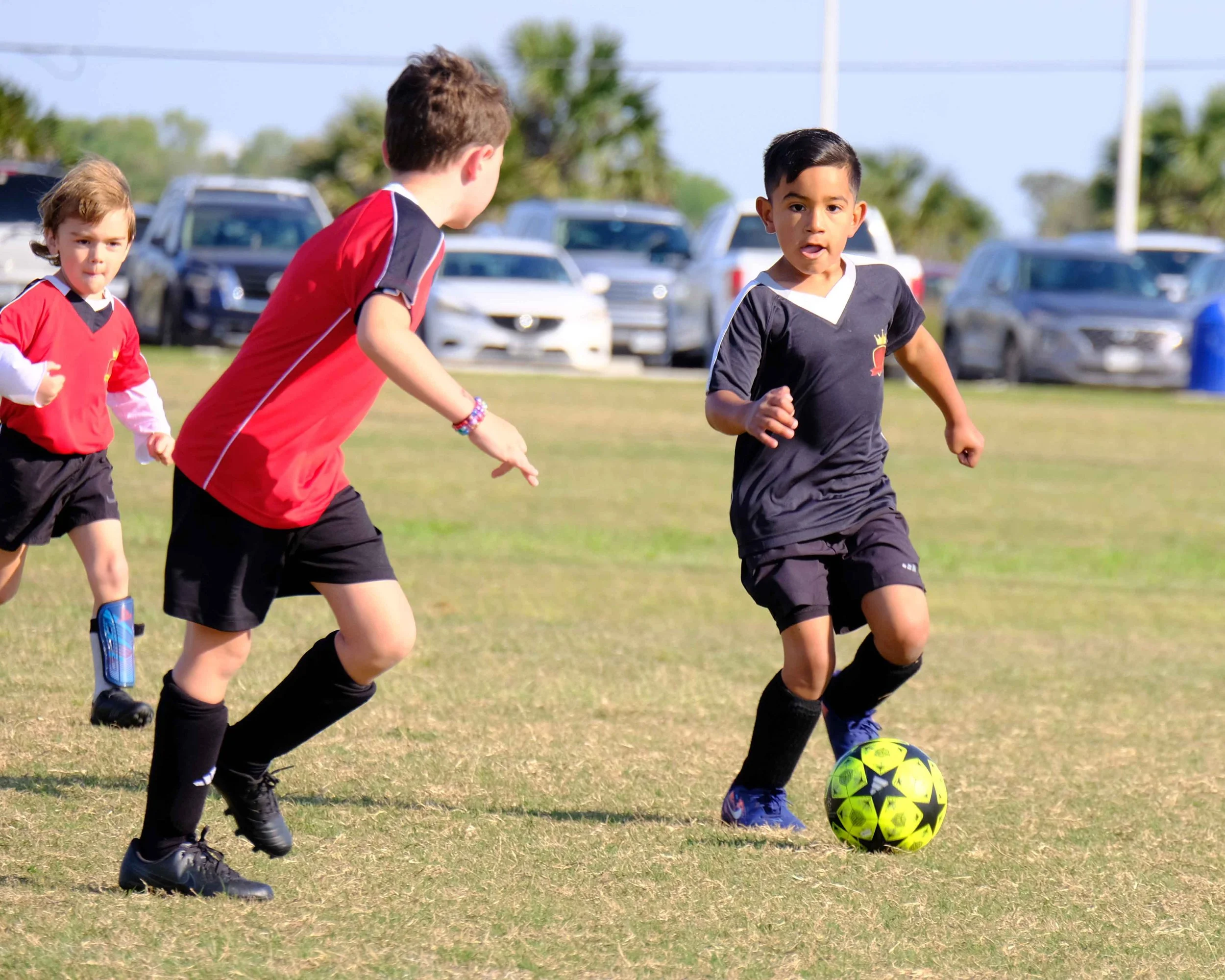 Young boys playing soccer on a grassy field, with a soccer ball at their feet and cars in the background.