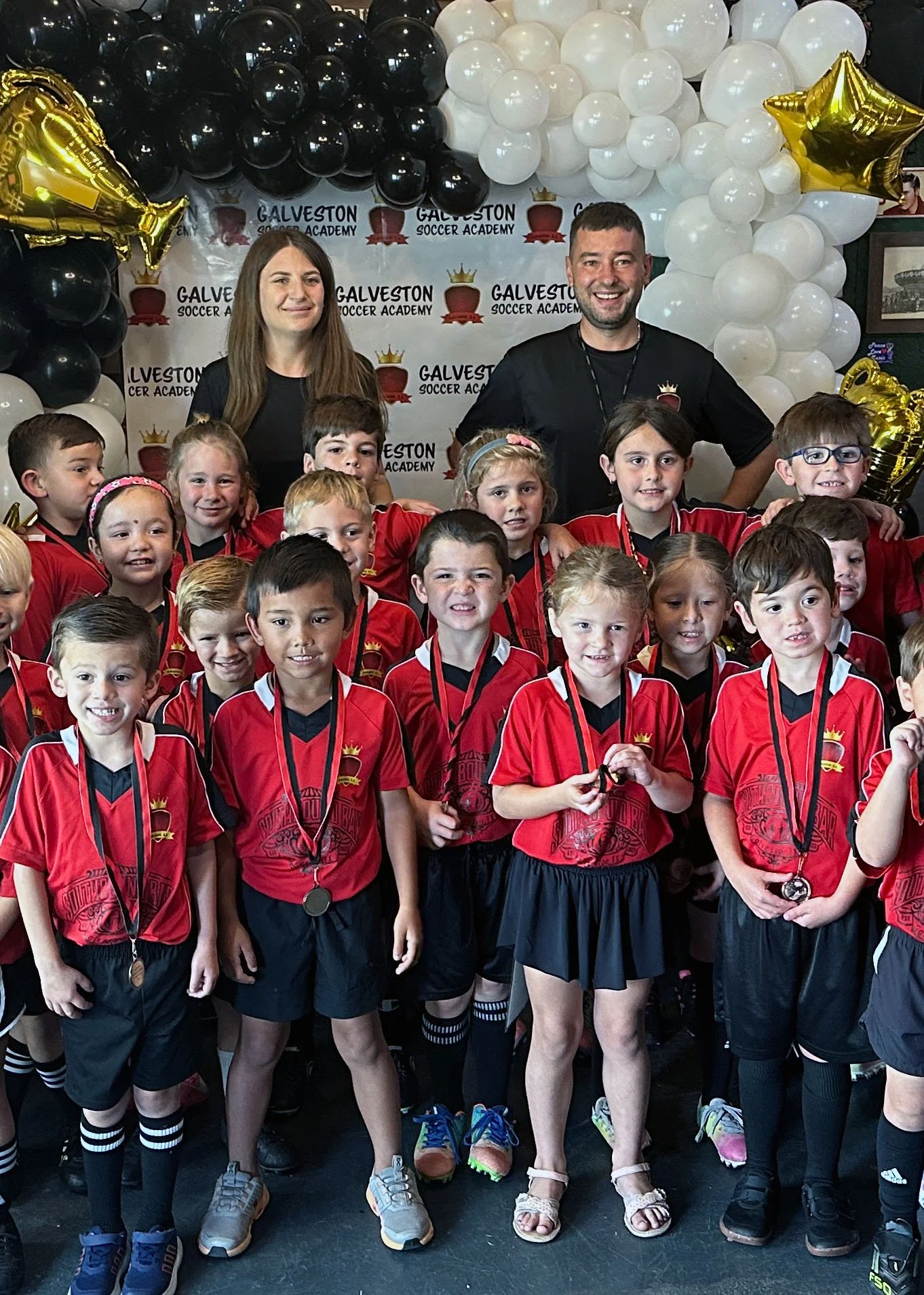 Group of young children wearing red soccer jerseys with medals around their necks, standing in front of a backdrop with balloons and the logo of Galveston Soccer Academy. Two adults stand behind the children, celebrating their achievement.