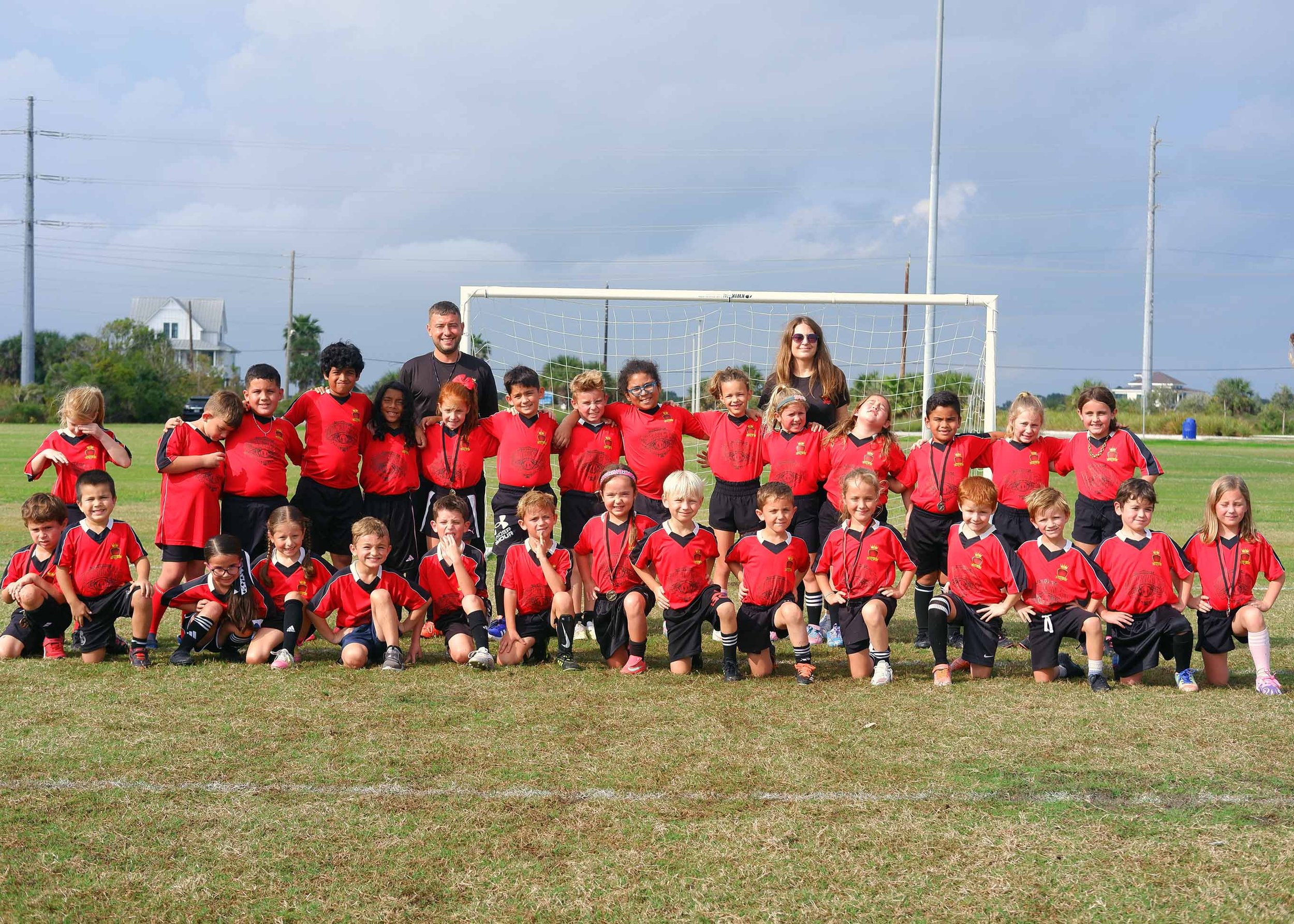 Group of children and two coaches posing on a soccer field with a goalpost in the background. The children wear red team uniforms and are smiling.