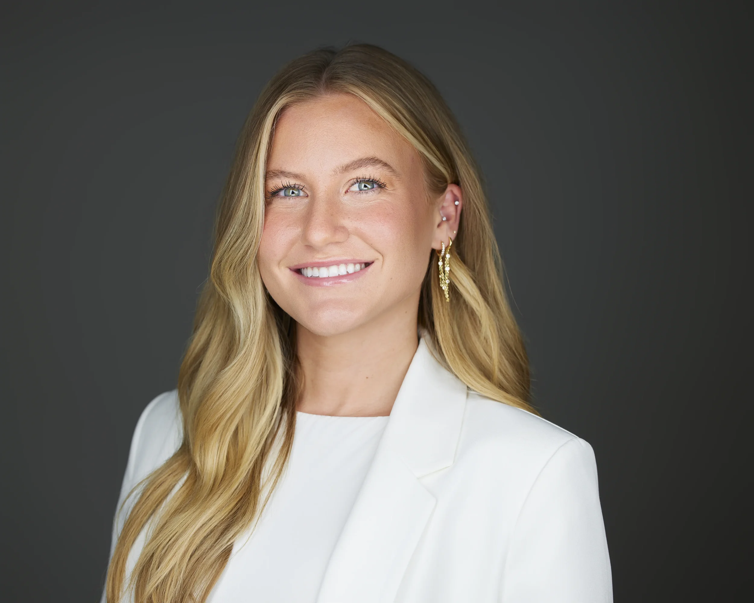 A woman with long blonde hair, wearing a white blazer and gold earrings, smiling in front of a dark background.