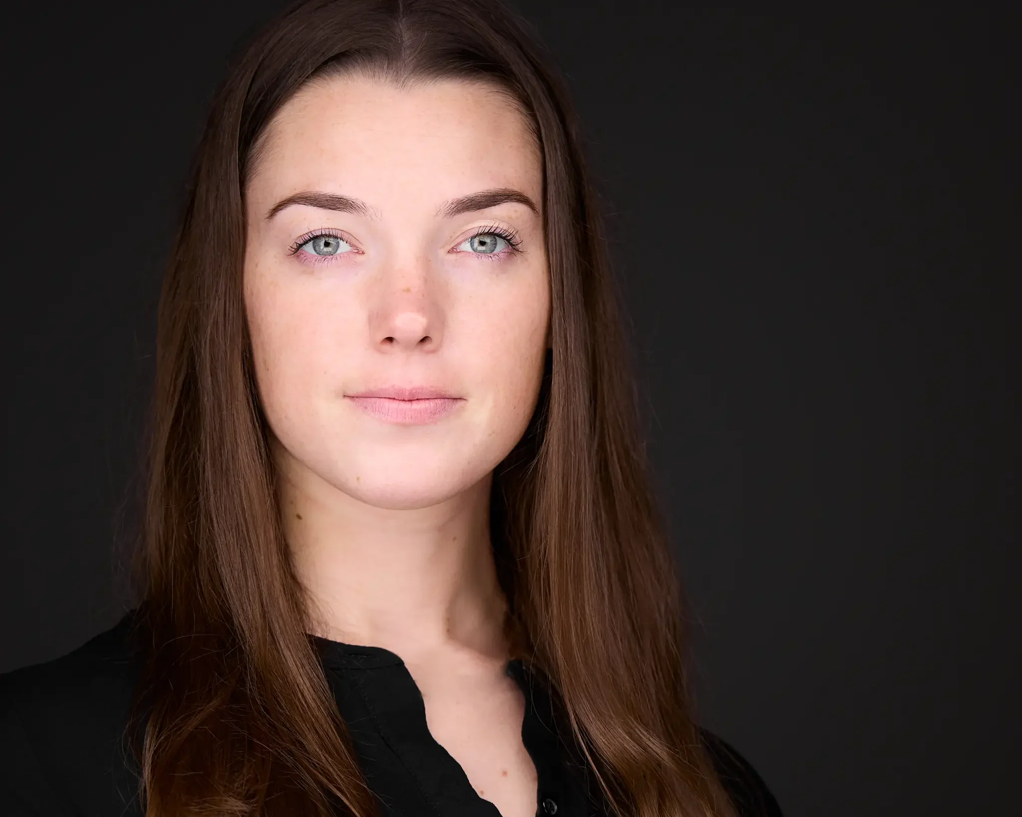 portrait of a young woman with long brown hair and light skin, wearing a black top, against a black background.