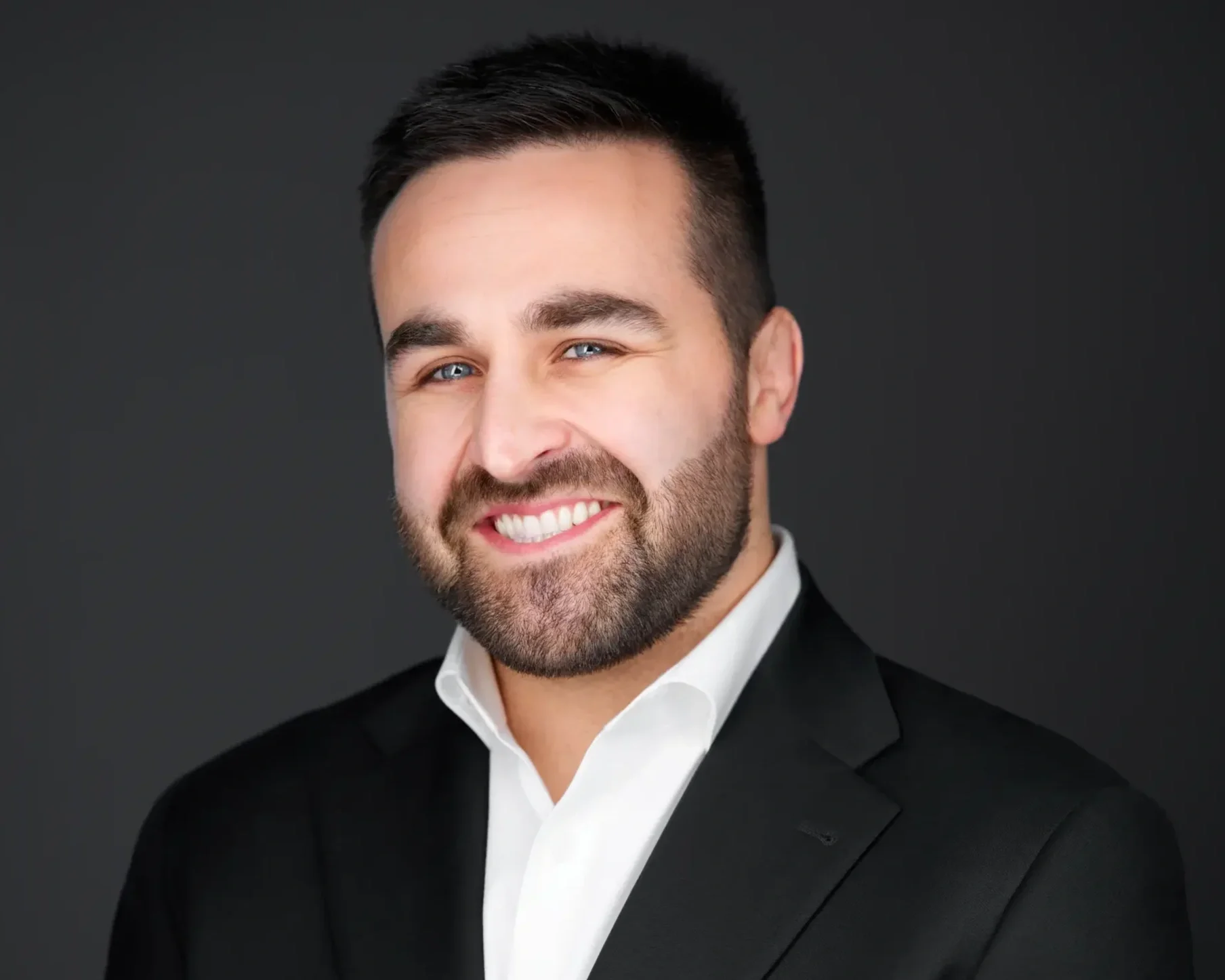 A smiling man with dark hair and a beard wearing a black suit and white shirt against a dark background.