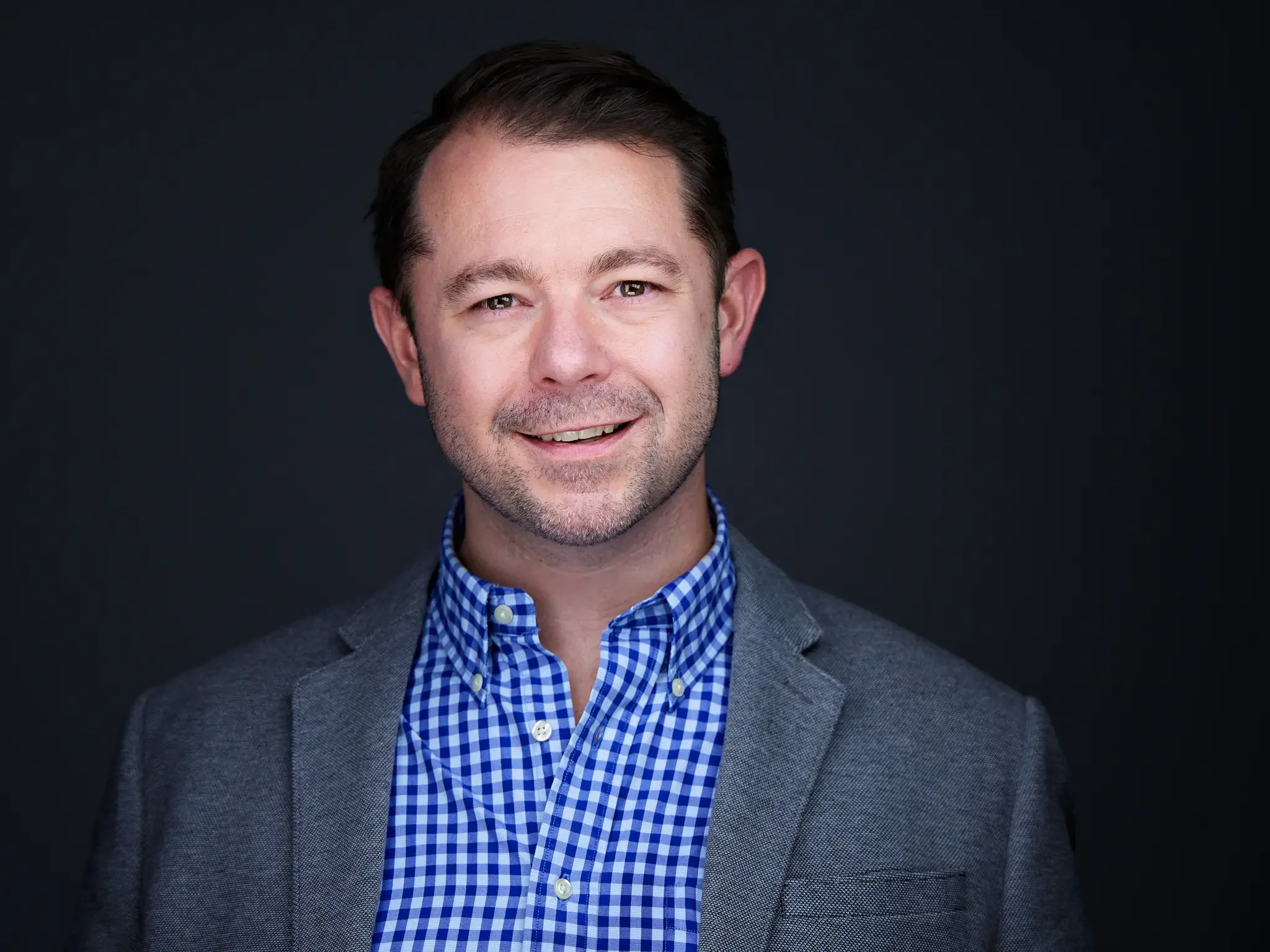 A man with brown hair and a beard smiling, wearing a blue checkered shirt and a gray blazer, against a dark plain background.