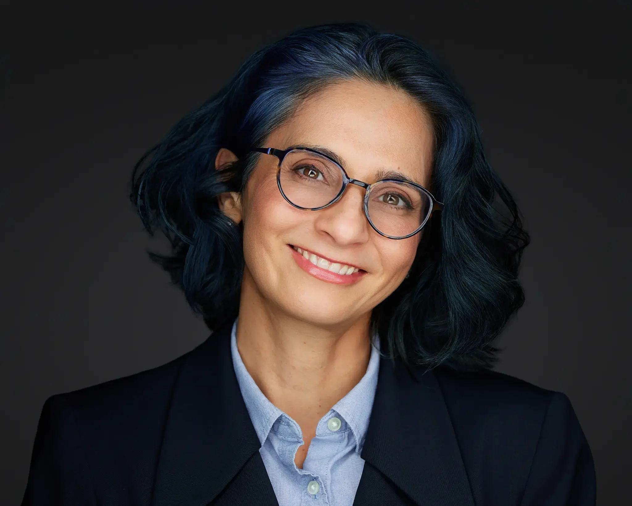 Portrait of a woman with shoulder-length dark hair and glasses, smiling, wearing a suit jacket over a light blue shirt against a dark background.