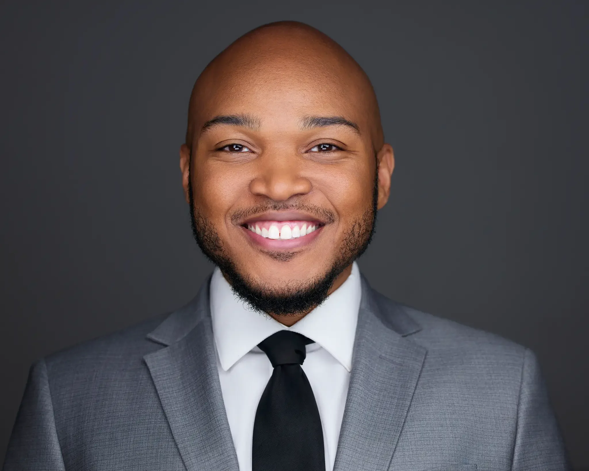 Professional portrait of a smiling Black man in a gray suit, white shirt, and black tie, against a dark background.