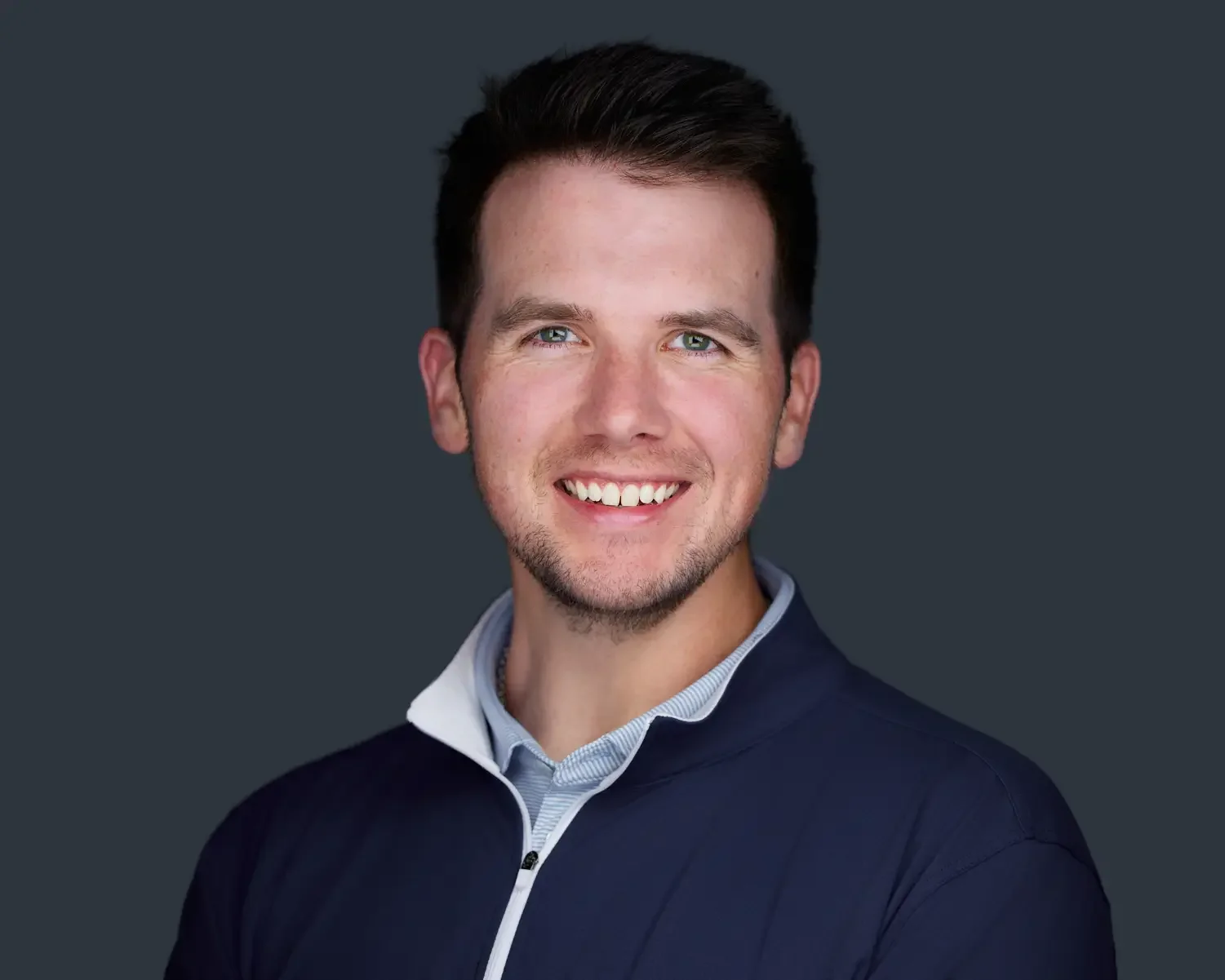 Portrait of a smiling young man with short dark hair, light skin, wearing a navy blue jacket over a light blue collared shirt, against a dark background.