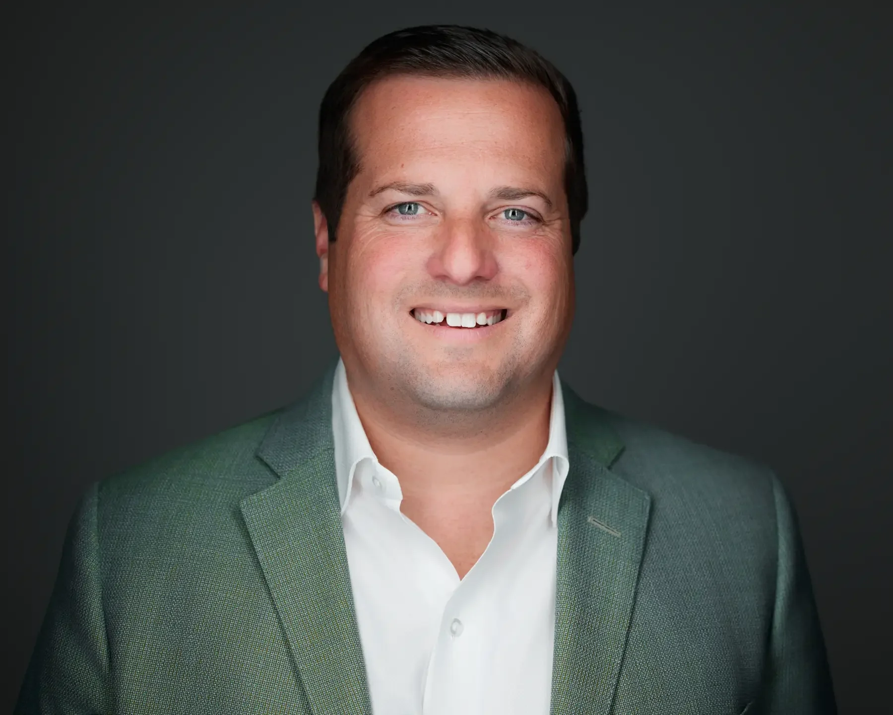 Headshot of a smiling man with dark hair, wearing a gray suit jacket and white dress shirt, against a dark gray background.