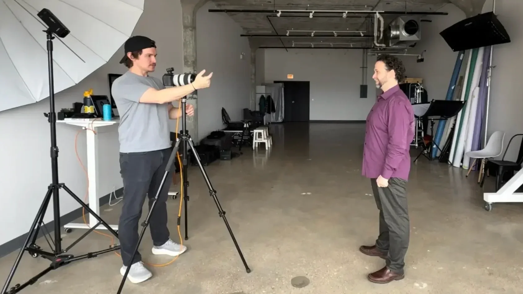 A man in a purple shirt standing in front of another man with a camera on a tripod in a photography studio.