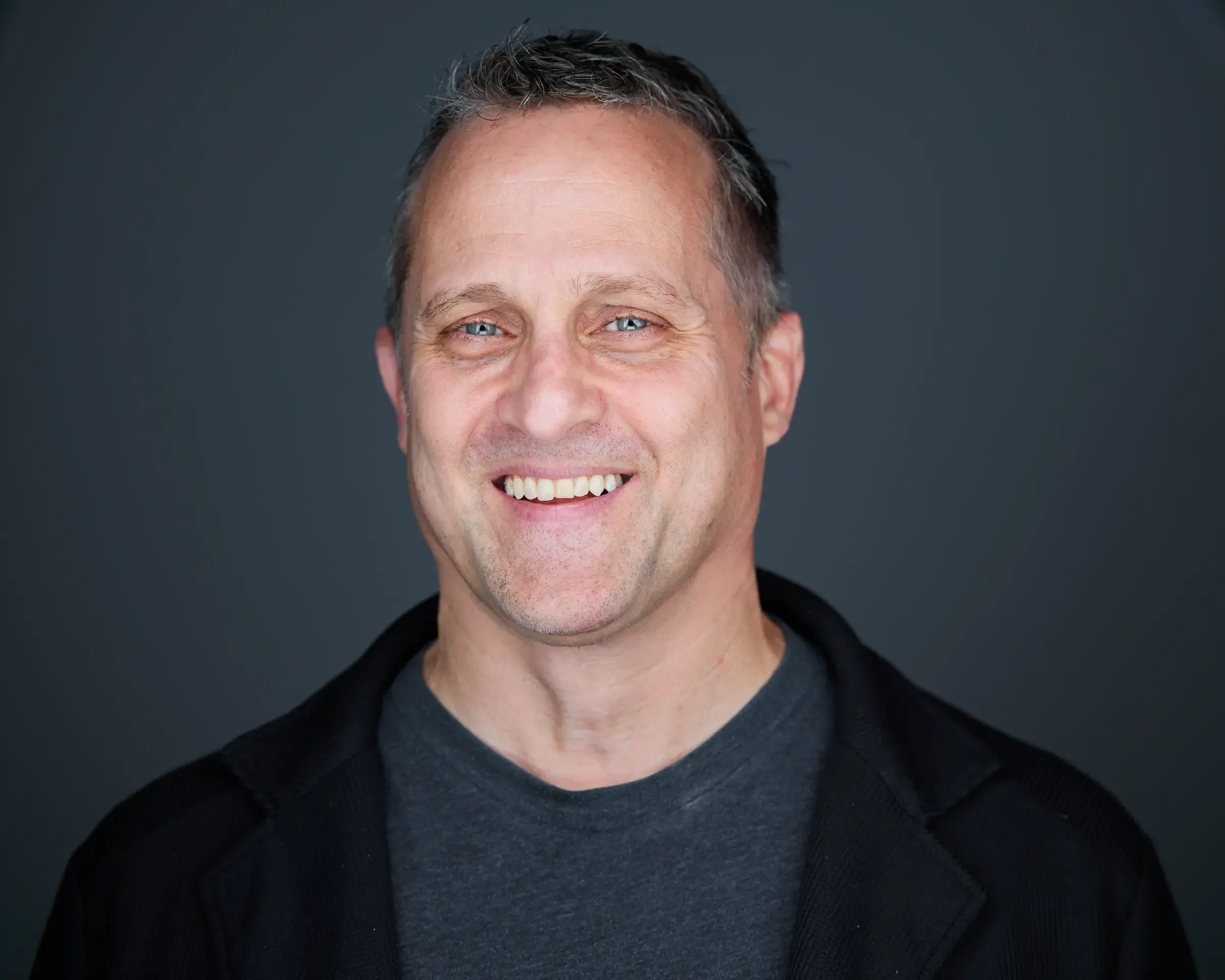 A smiling man with short, slightly tousled gray hair, blue eyes, and light skin, wearing a dark jacket over a gray t-shirt, standing against a dark background.