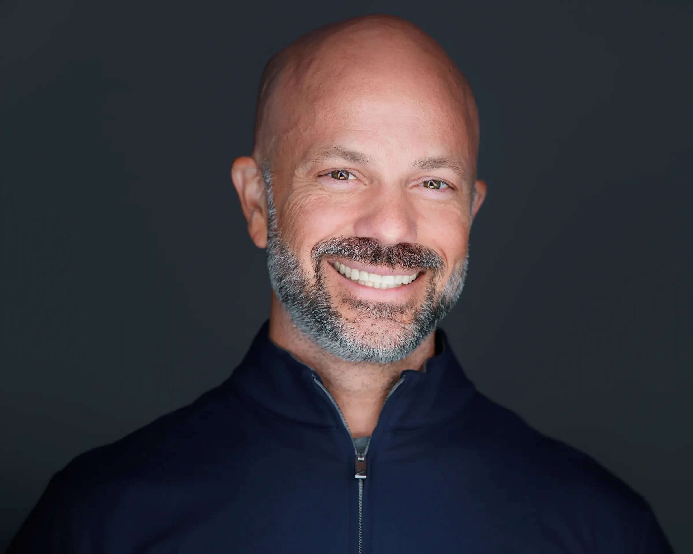 Headshot of a smiling middle-aged man with a beard and bald head, wearing a dark zip-up jacket, against a dark background.