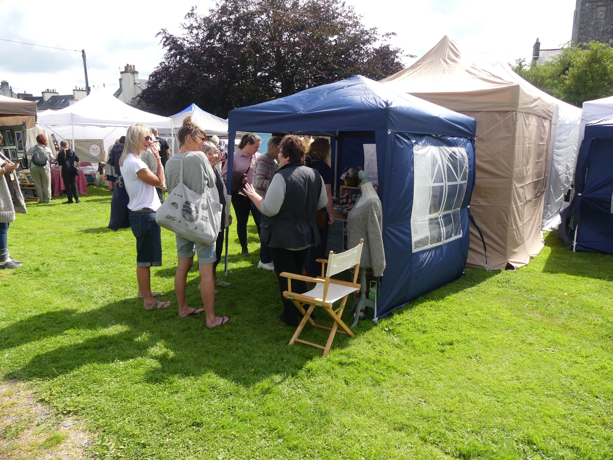 People browsing market stalls at an outdoor fair on grass, with tents and trees in the background.