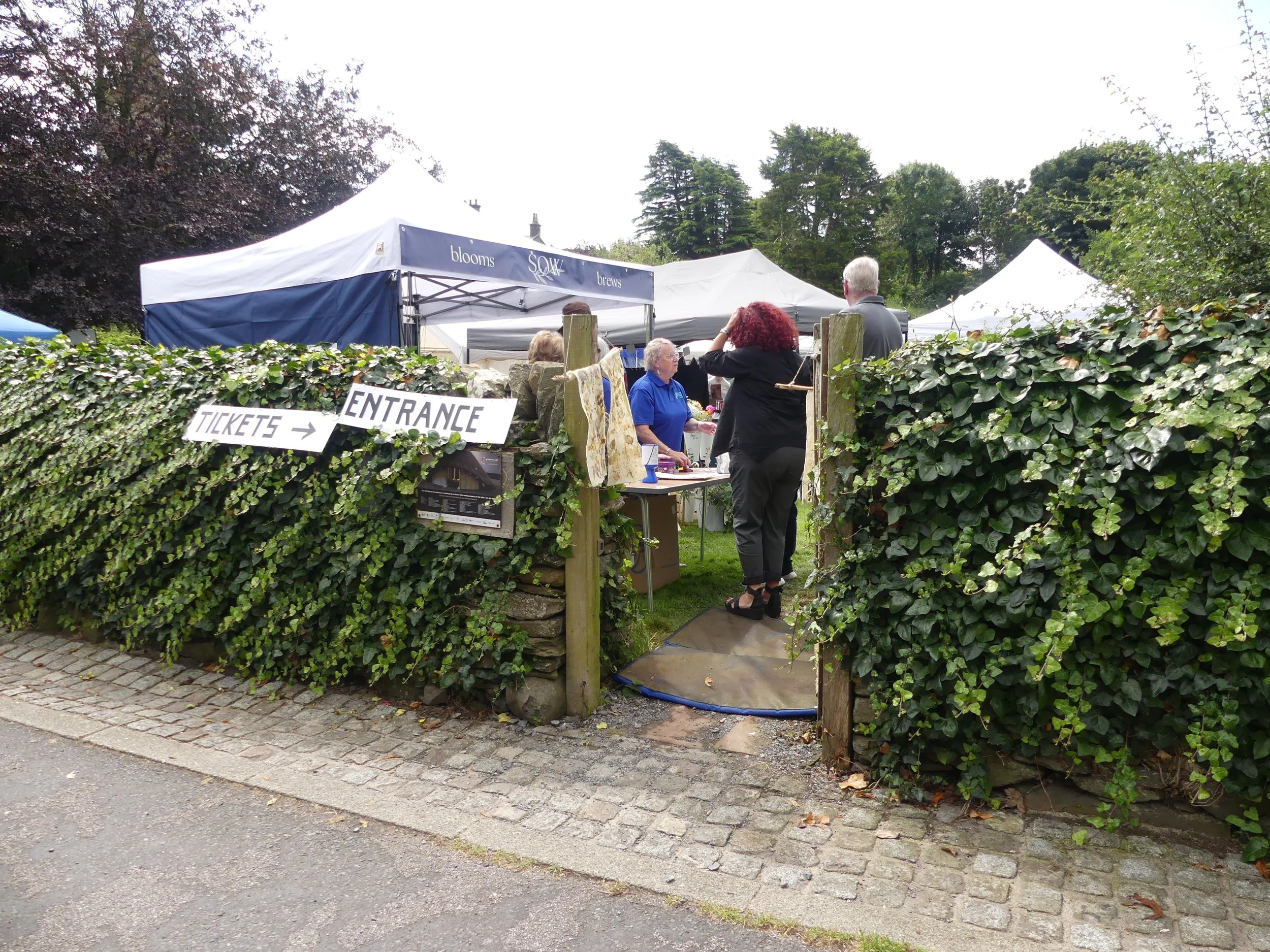 People at an outdoor market entrance with tents, signs, and a hedge