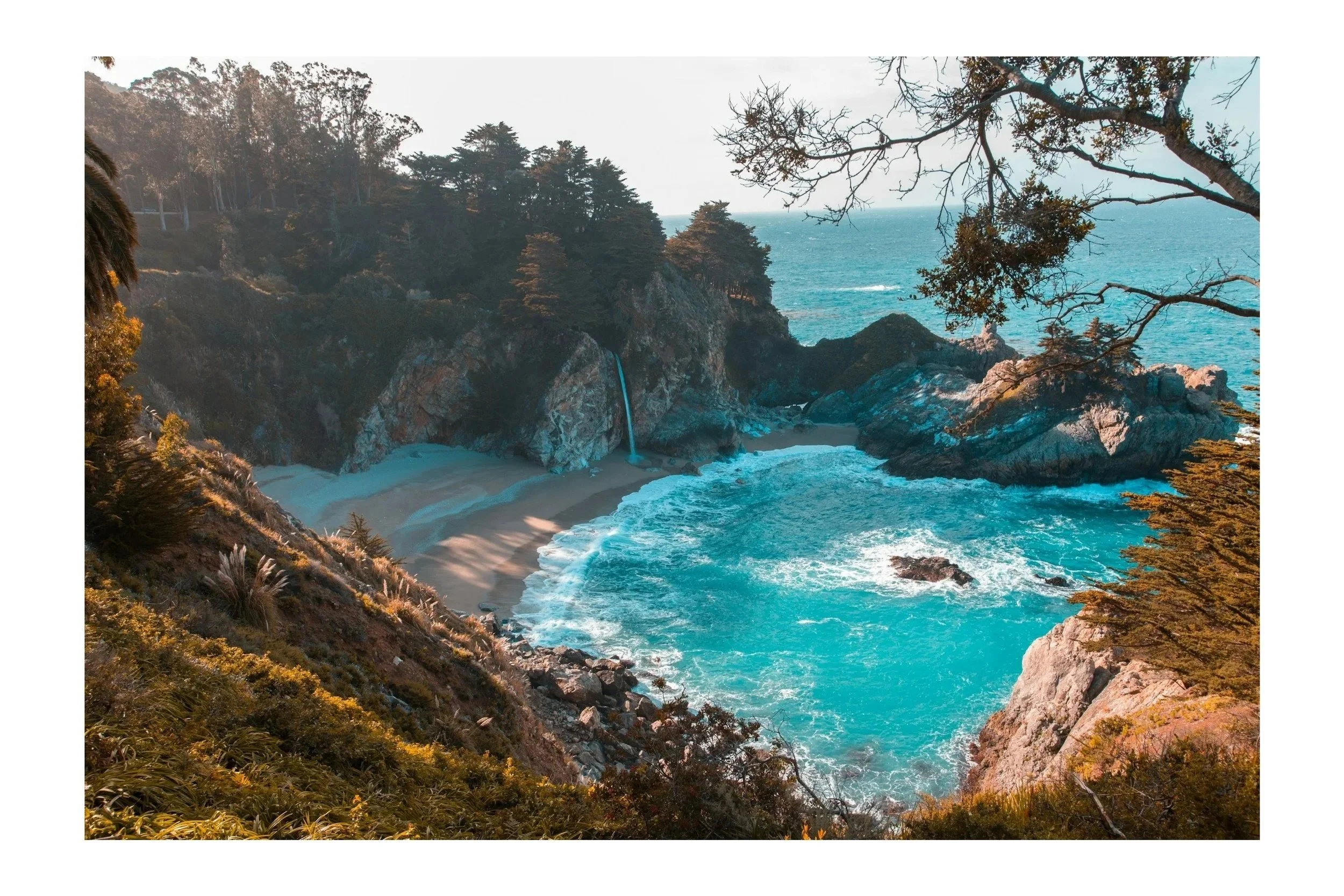 A scenic coastal view of a cove with a beach, turquoise water, rocky cliffs, a waterfall flowing into the ocean, surrounded by trees and foliage, under a clear sky.