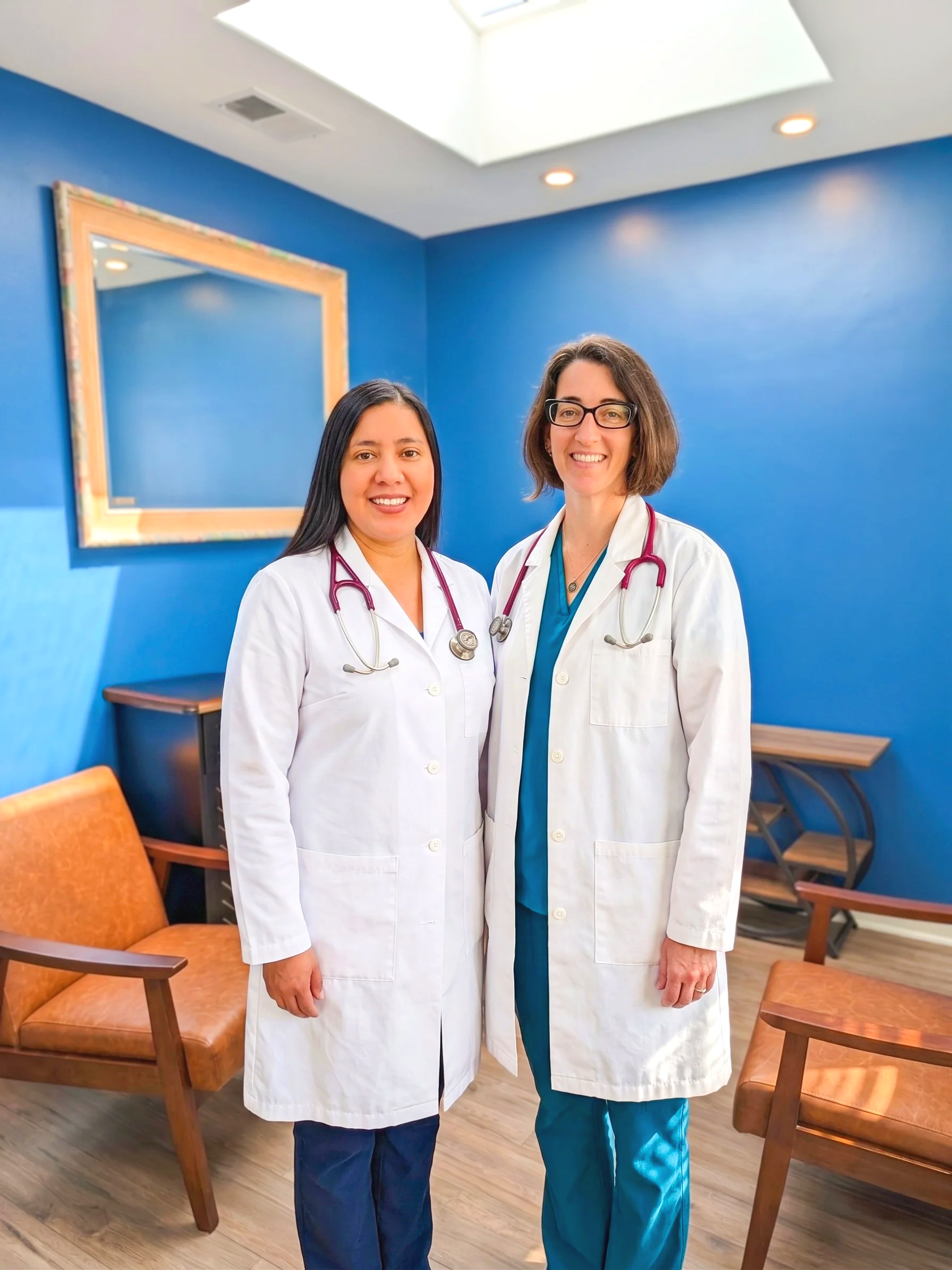 Two female doctors standing together in a medical office, wearing white lab coats and stethoscopes, smiling at the camera.