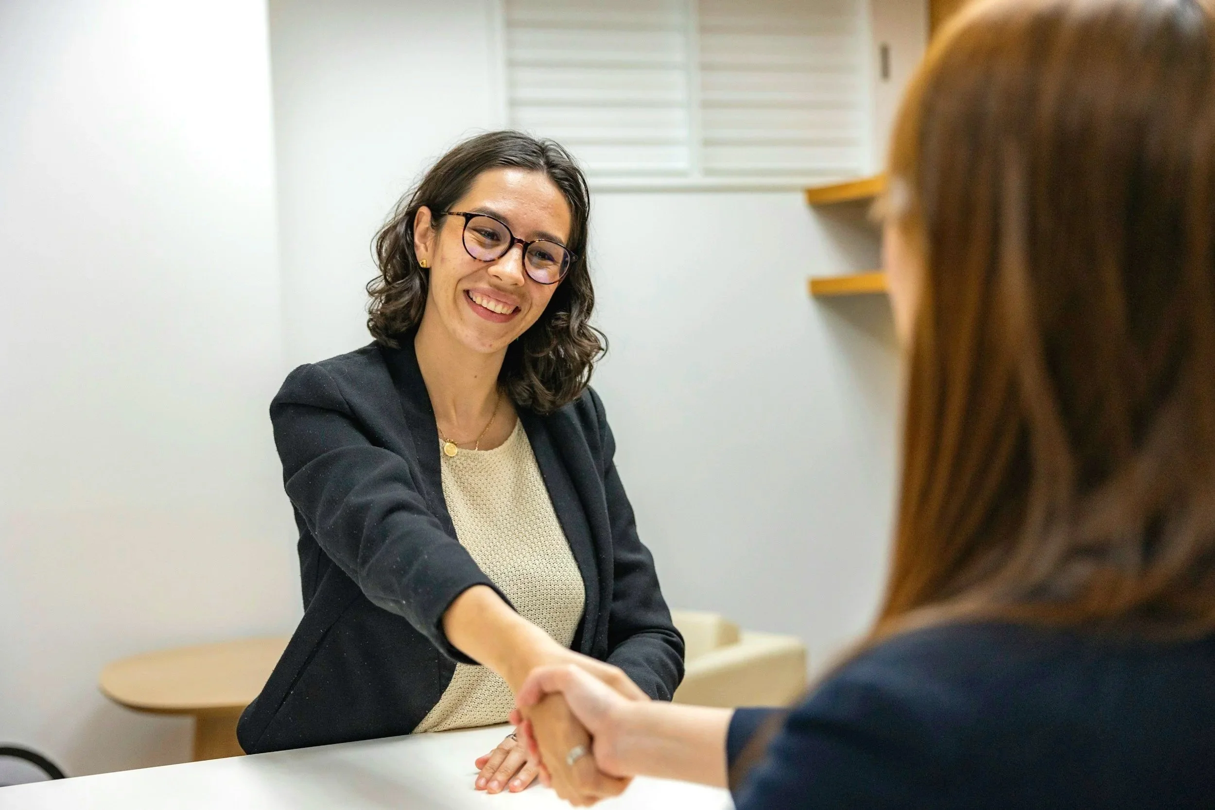 Two women shaking hands in an office, one smiling and wearing glasses, the other with red hair, with shelves on the wall behind them.