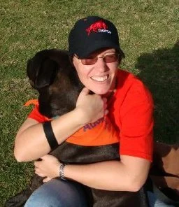 A woman smiling and wearing sunglasses, a black cap, and a red and orange shirt, hugging a black dog outdoors on grass.