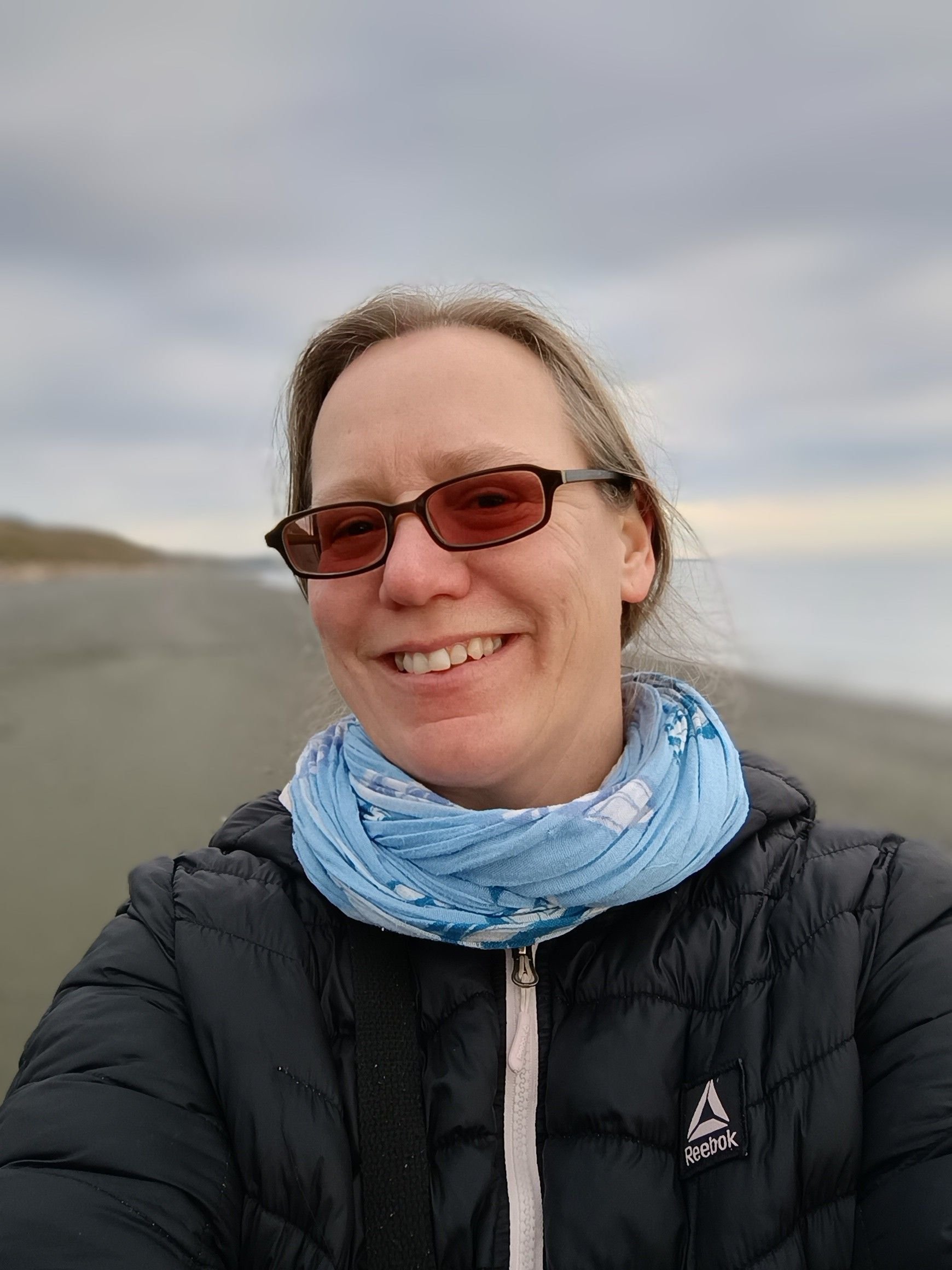Smiling woman with blonde hair wearing sunglasses, a blue scarf, and a black puffer jacket outside, with a cloudy sky in the background.
