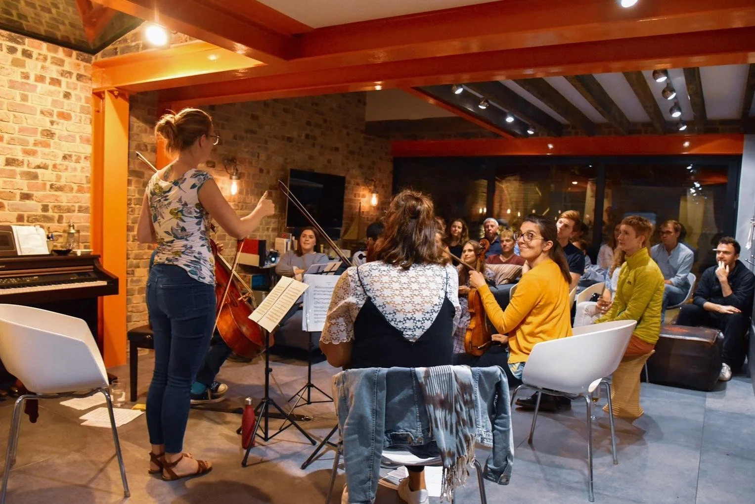 Florian Ensemble violist stands with her instrument, addressing the small audience group in a cozy house concert. Brick walls and warm lighting, with audience seated and listening attentively.