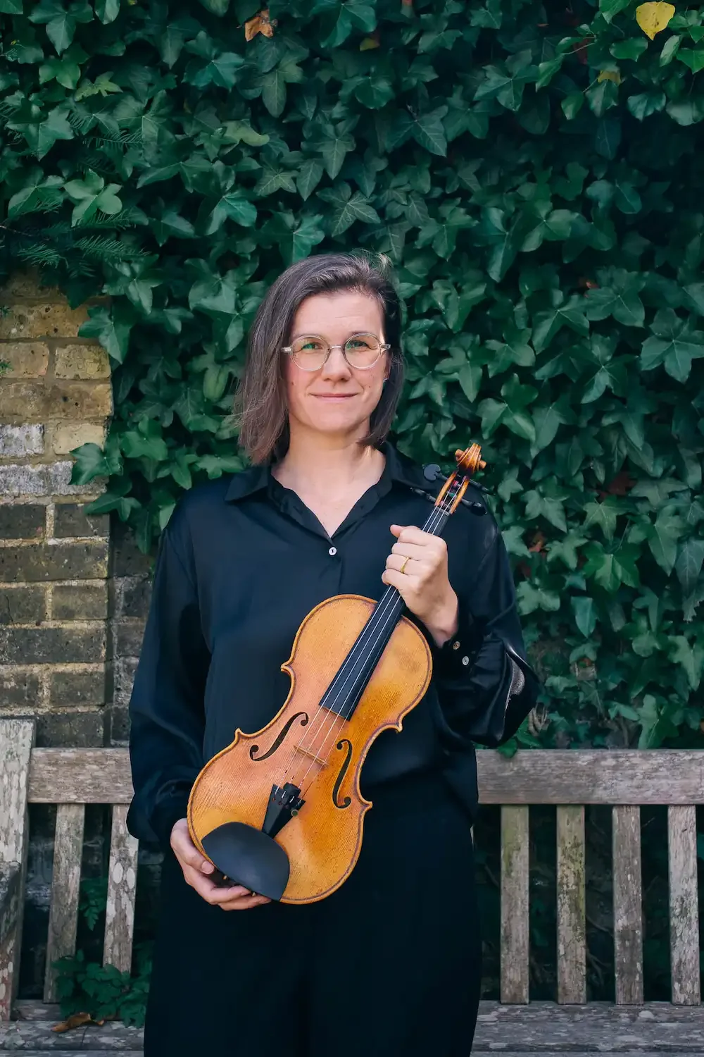 Violinist Kay posing with her instrument in front of a bench and greenery