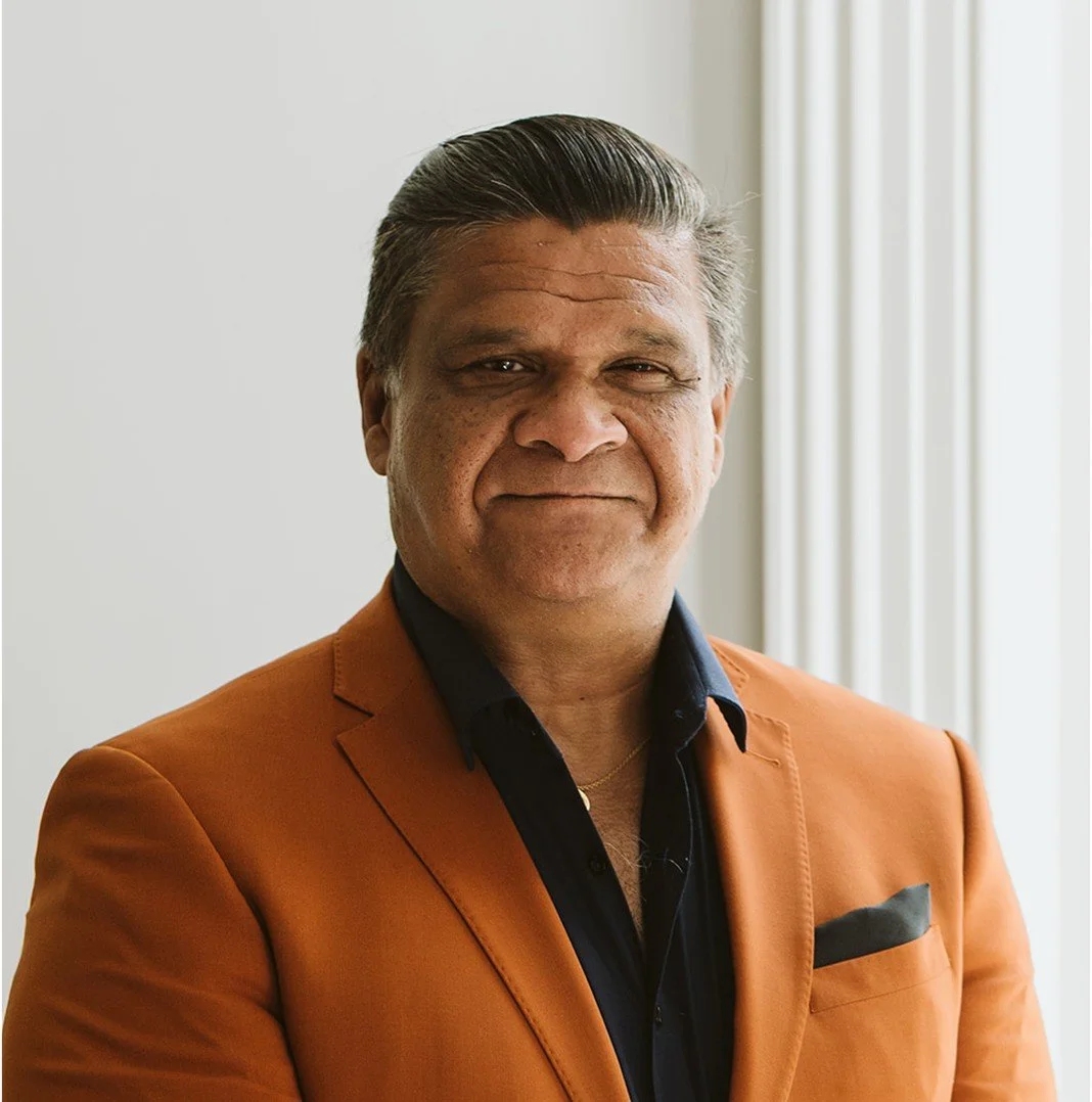 A middle-aged man in an orange blazer and black shirt standing indoors near a window with blinds.