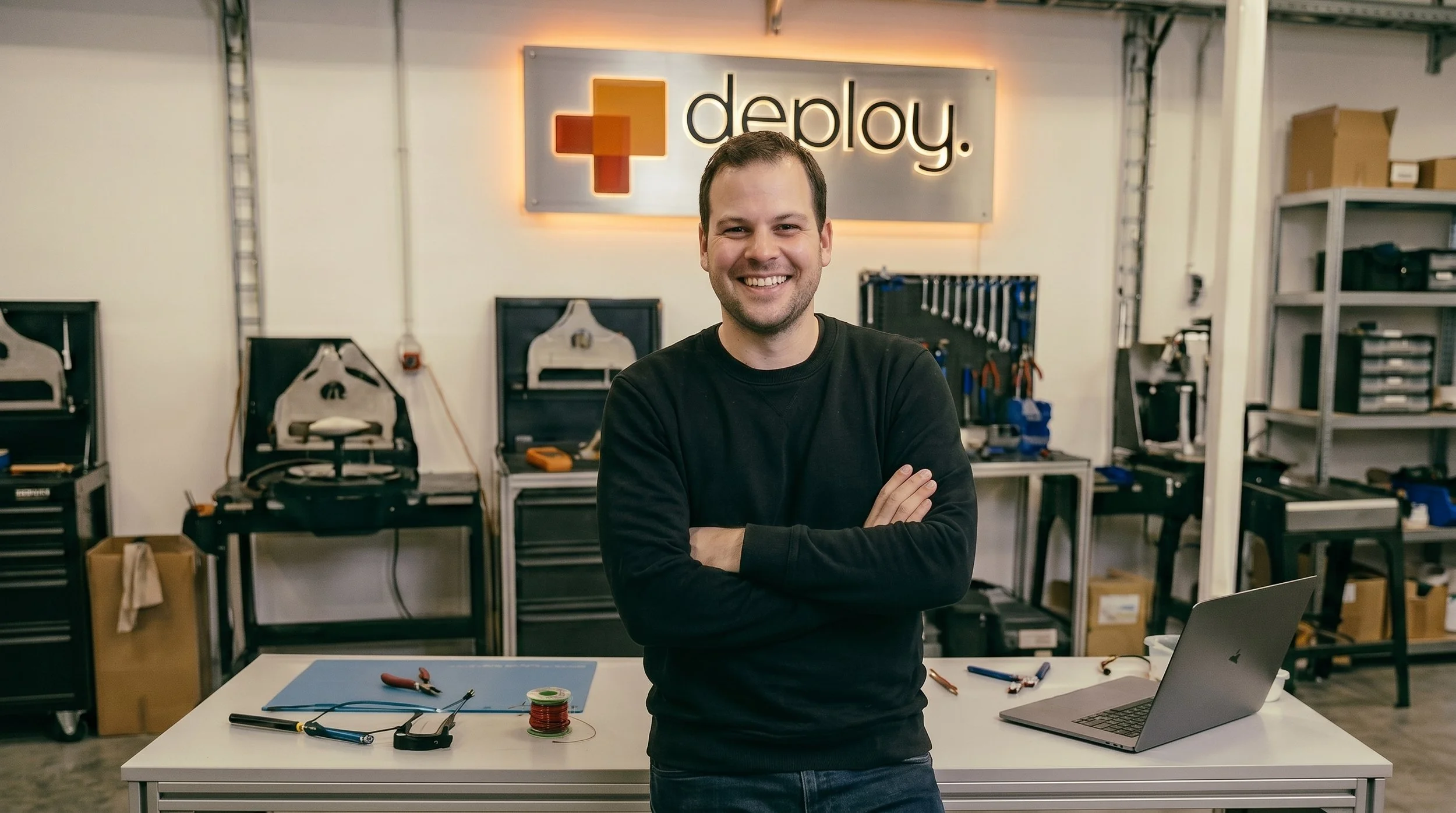A smiling man stands in a workshop or lab with arms crossed, in front of a workbench with tools, electronic components, and a laptop. Behind him is a sign that says 'deplay' with a red and orange geometric shape.