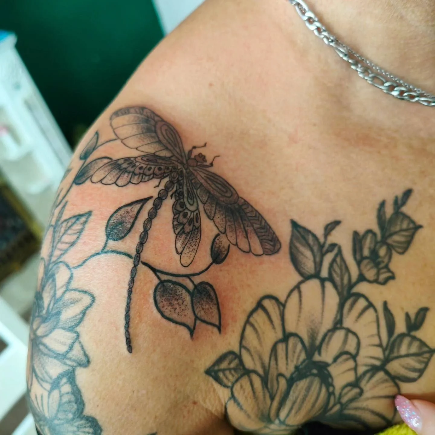 Close-up of a tattoo on a person's shoulder and upper chest, featuring a detailed black and gray dragonfly and floral design.