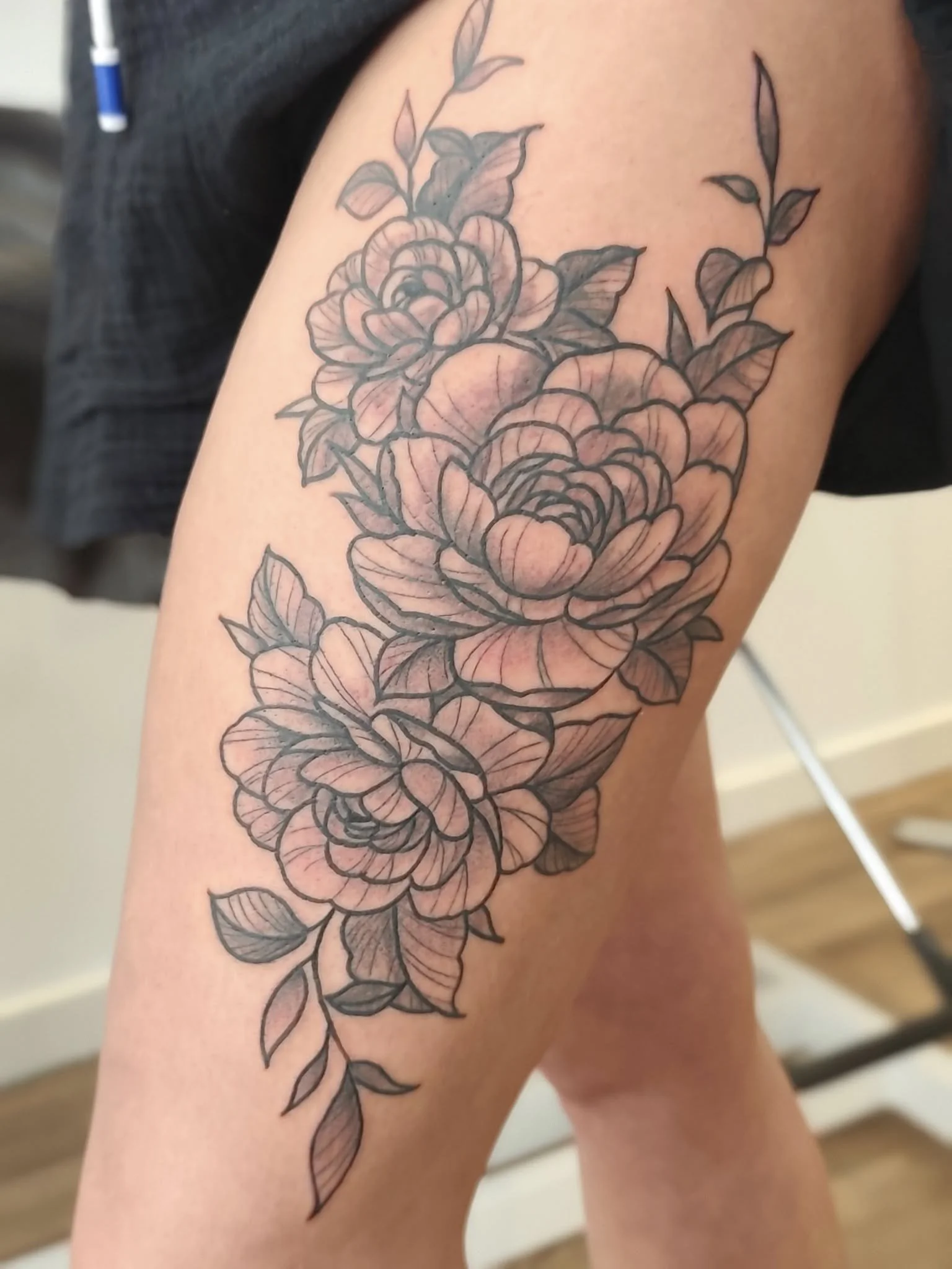 Black and gray floral tattoo on person's thigh featuring multiple peonies and leaves.