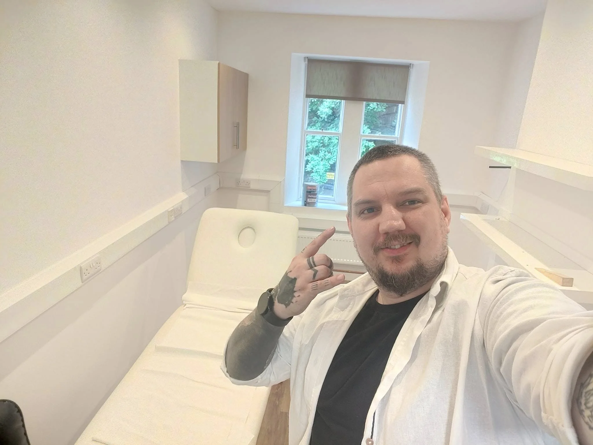 A man taking a selfie in a tattoo studio room with a white bed and a window showing greenery outside.