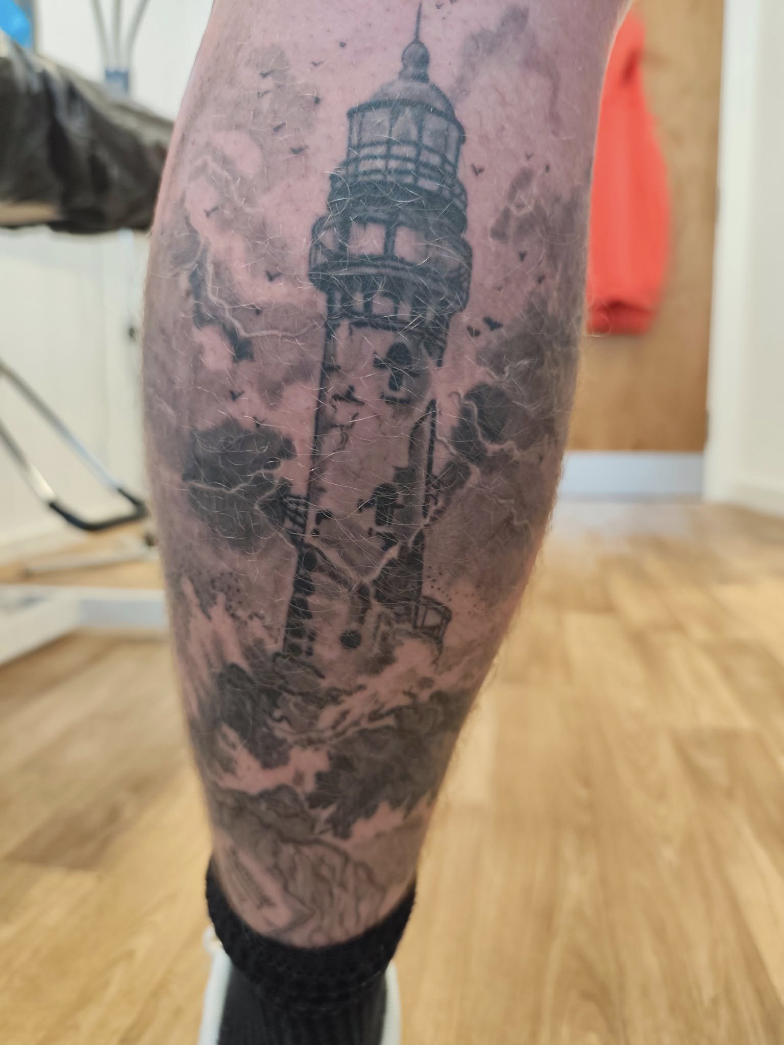 Tattoo of a lighthouse on a person's thigh, with clouds during a storm in the background.