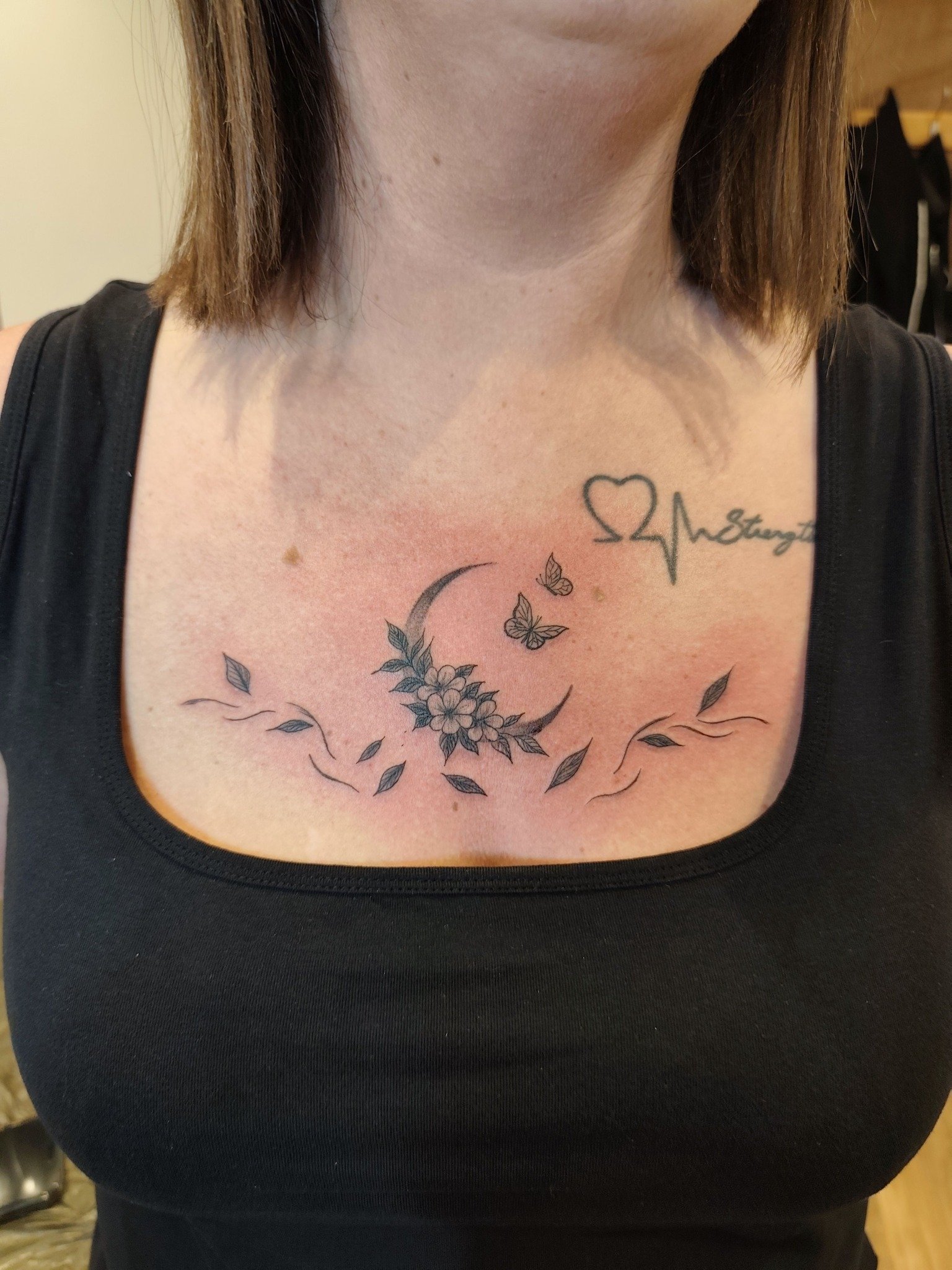 Tattoo on woman's chest featuring a crescent moon with flowers, butterflies, and leaves, with a heart and heartbeat line with text above.