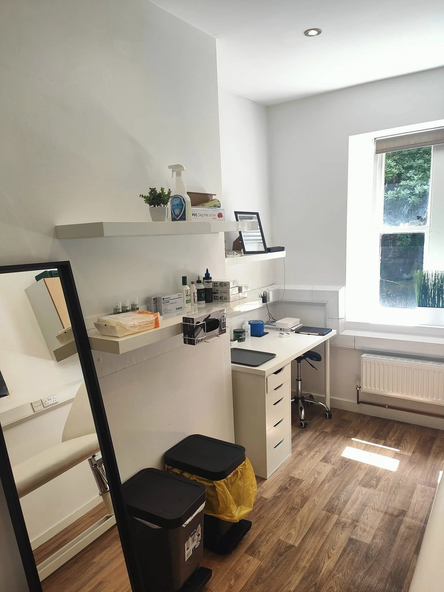 A small, tidy tattoo studio office room with a white desk, a rolling chair, and shelves with medical supplies. There is a window letting in natural light and a mirror leaning against the wall. Trash bins are visible near the mirror.