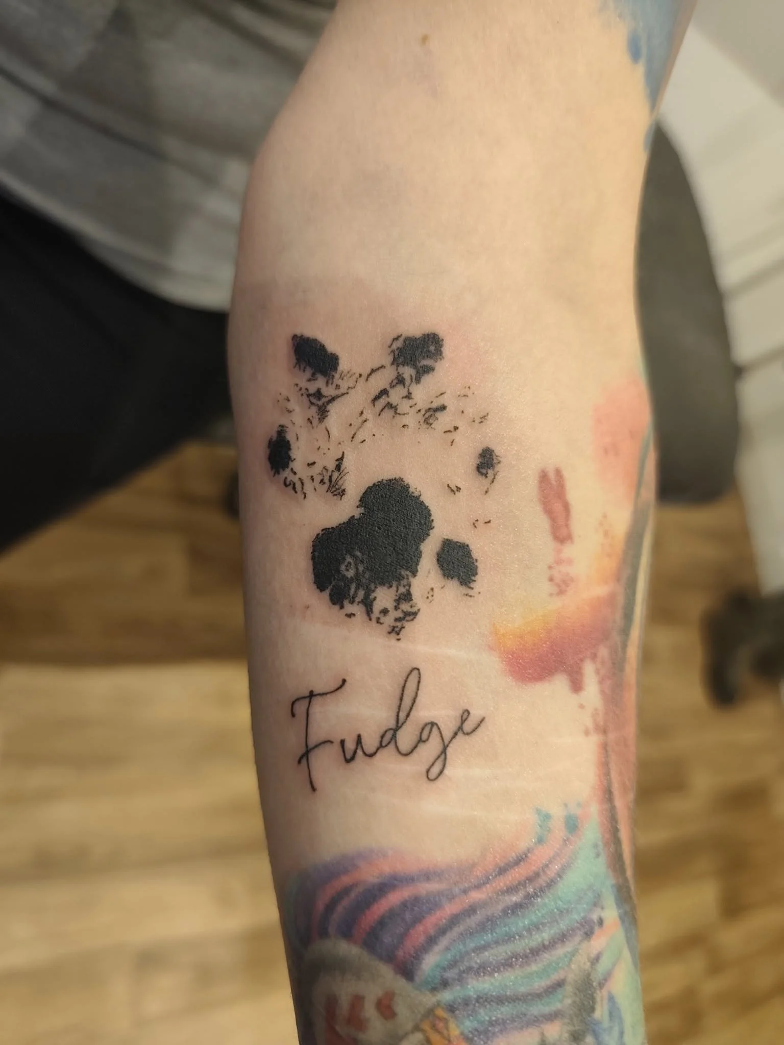 Tattoo of a dogs paw and the word 'Fudge' written in cursive on a person's arm.
