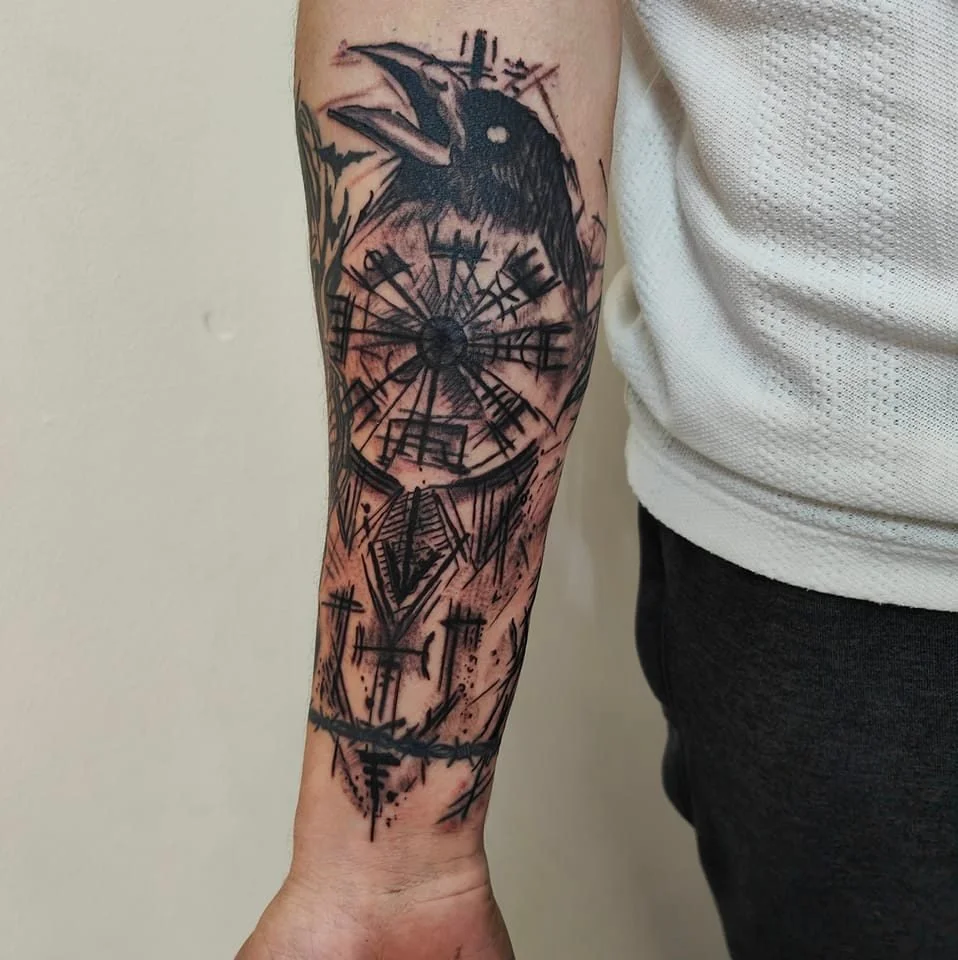 Black tattoo of a crow with a Nordic design and barbed wire on a person's forearm.